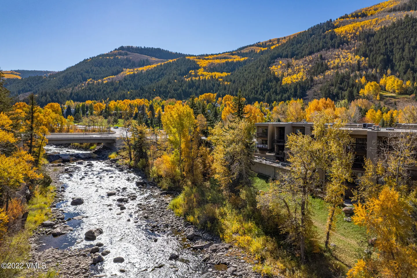 Aerial view of a rocky river flowing past multi-story residential buildings with vibrant autumn trees and forested mountains in the background.
