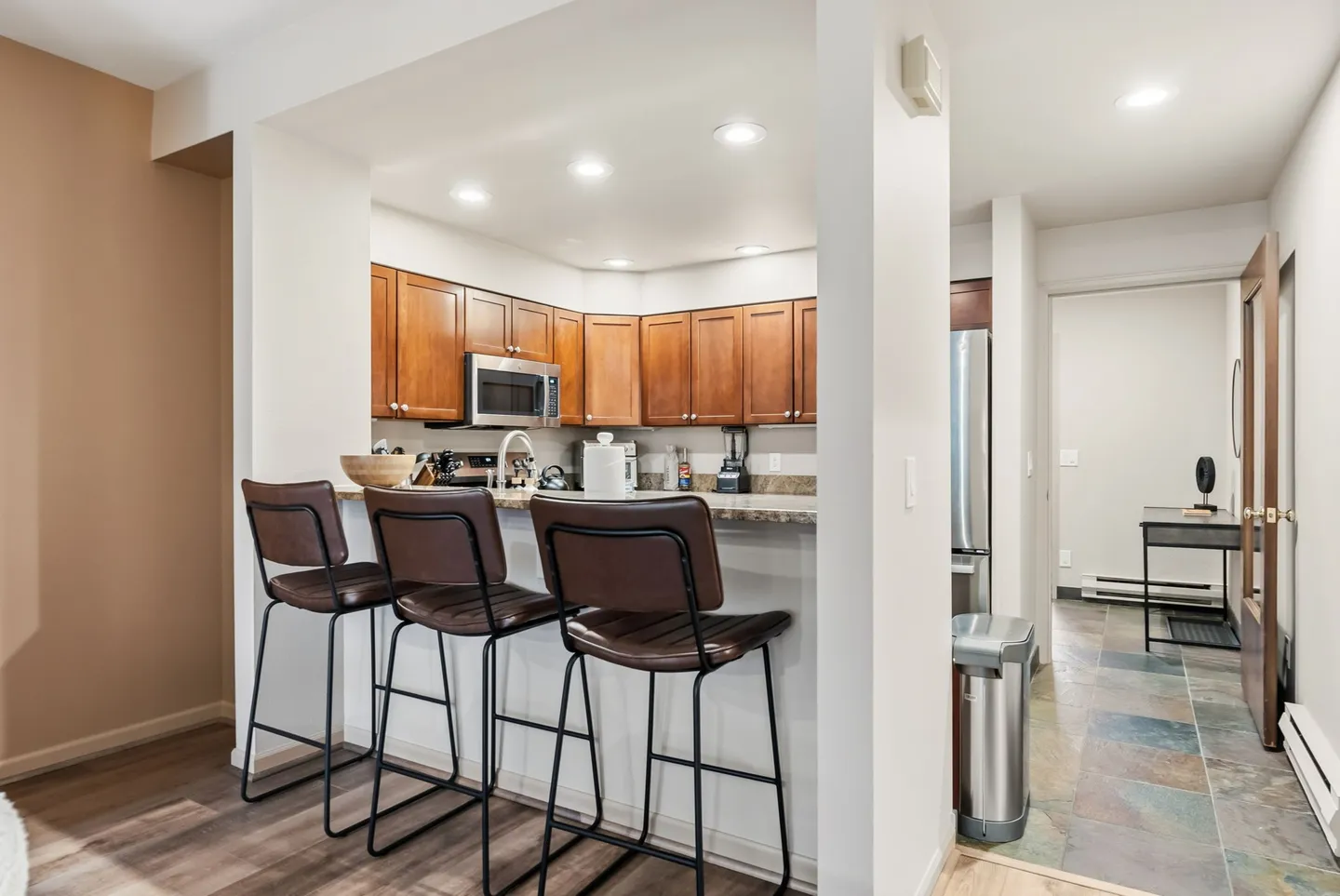 Kitchen counter with three modern bar stools, framed by wooden cabinets and recessed ceiling lights.