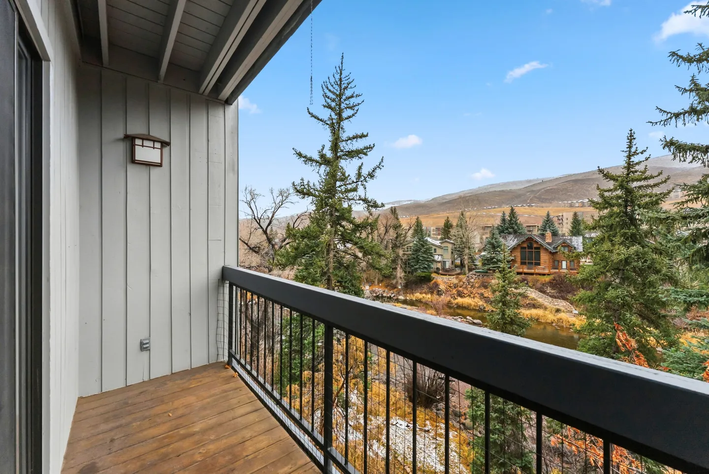 A covered balcony with a black metal railing overlooking pine trees, a waterway, and distant hills.