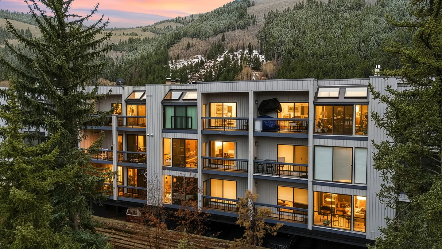 Exterior dusk view of a multi-story condominium building with lit balconies against a forested mountain.