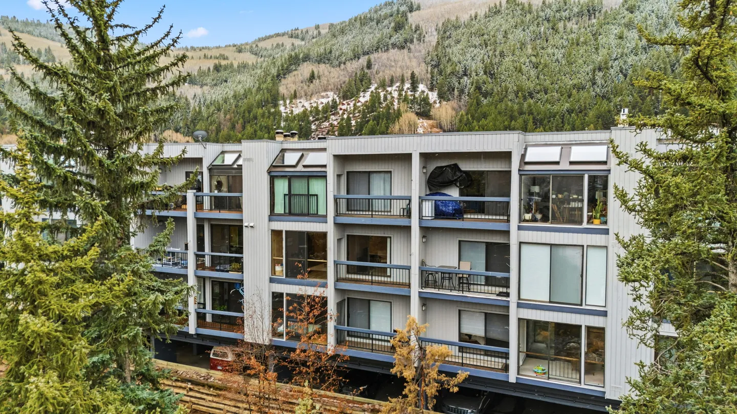 Exterior view of a multi-story condominium building with balconies, framed by pine trees against a mountain backdrop.