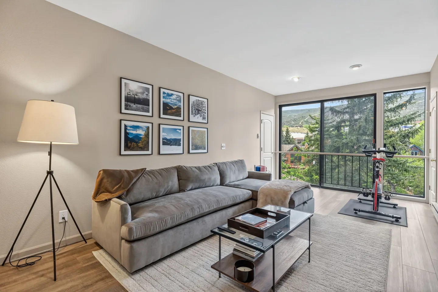 Living room featuring a grey sectional sofa, coffee table, tripod floor lamp, and a stationary exercise bike by large sliding glass doors.