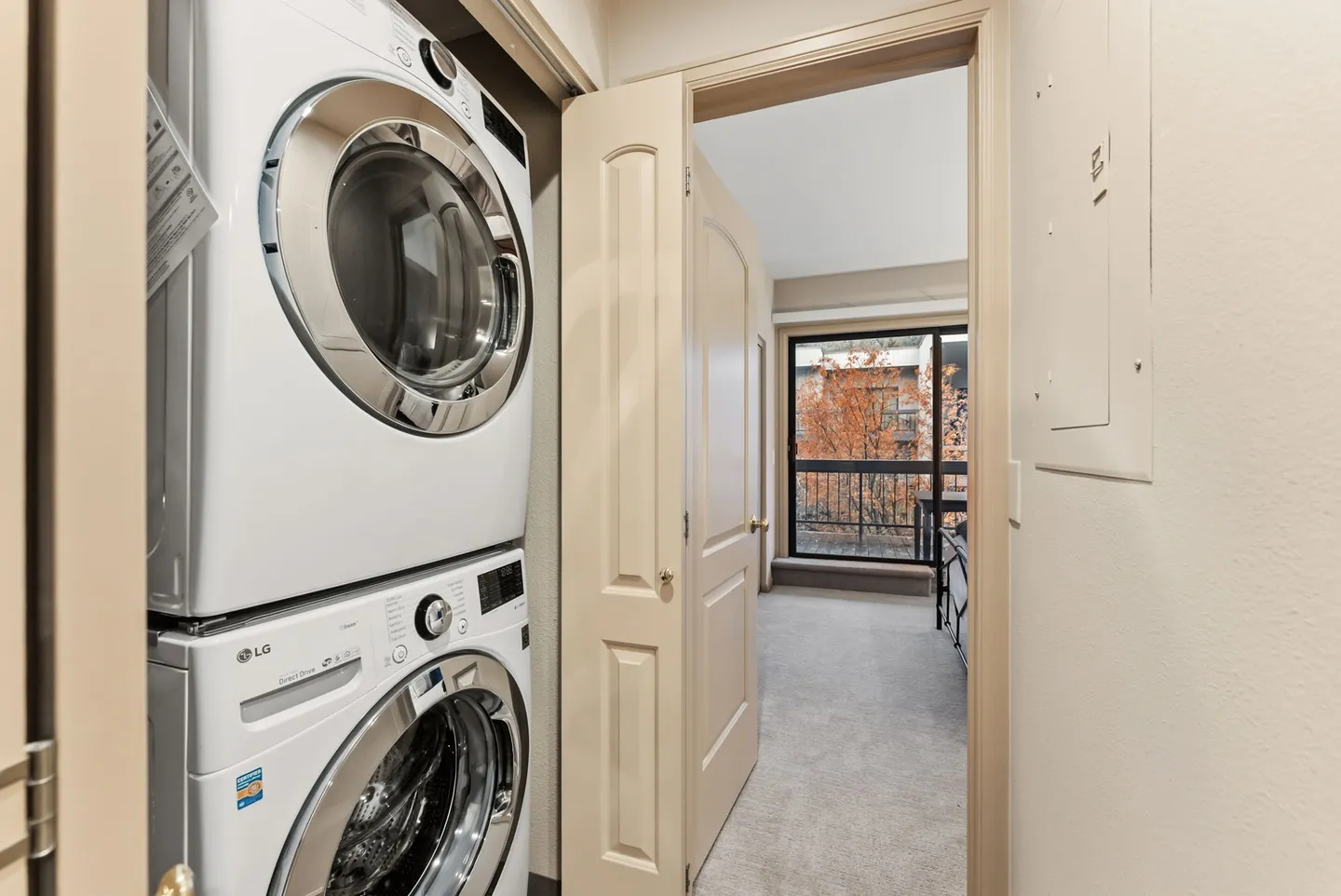 Stacked front-loading washer and dryer in a closet doorway leading to a carpeted bedroom with a sliding glass door.