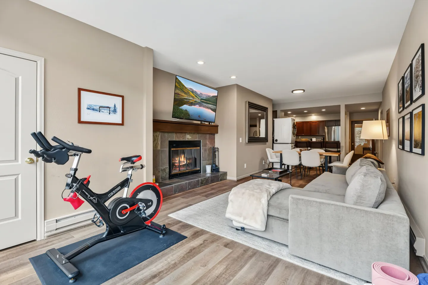 Open living area featuring a fireplace, mounted TV, sectional sofa, stationary bike, and dining space in the background.
