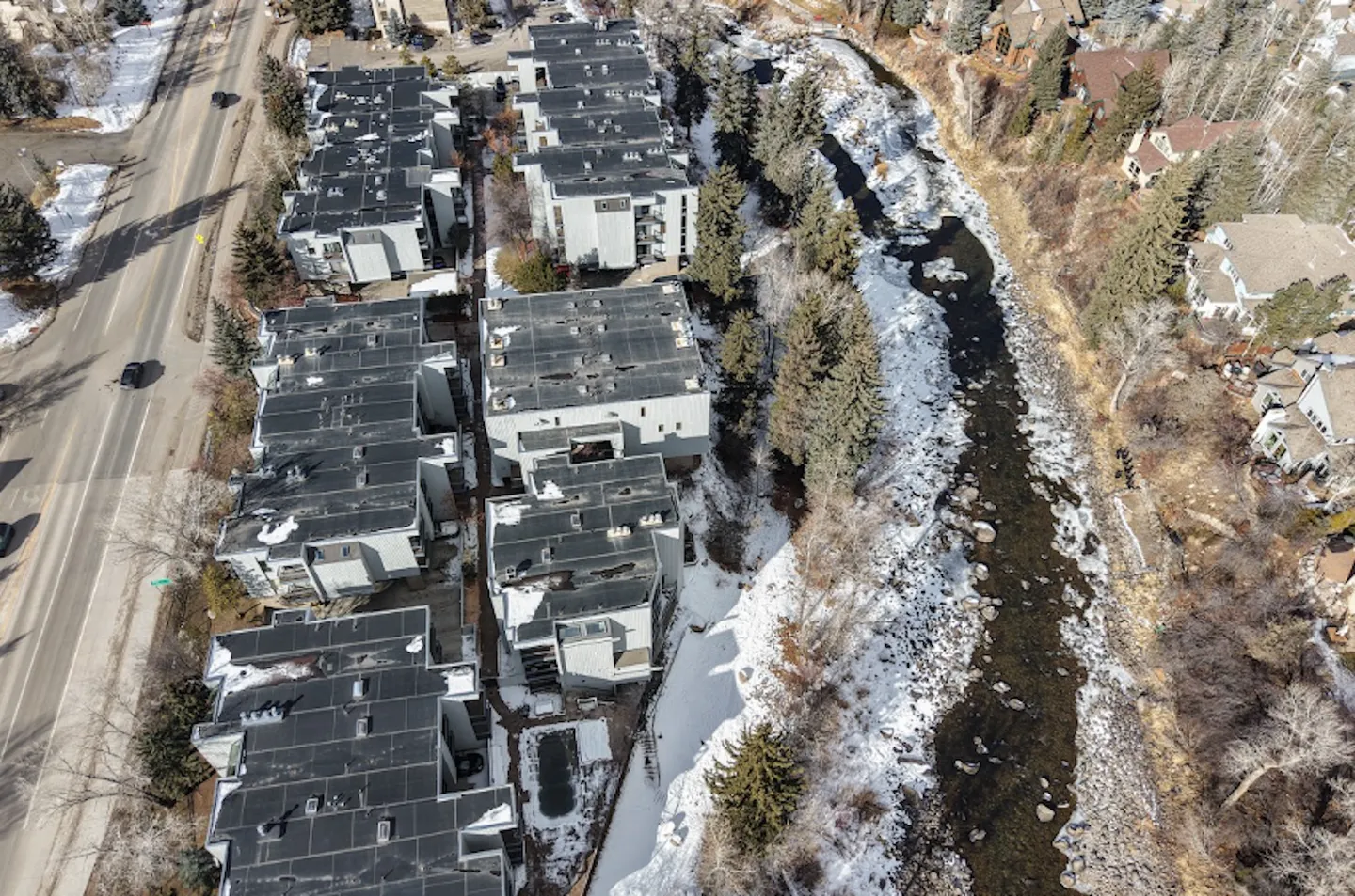 Aerial view of a multi-unit residential complex bordered by a roadway on one side and a rocky, snow-dusted river on the other.