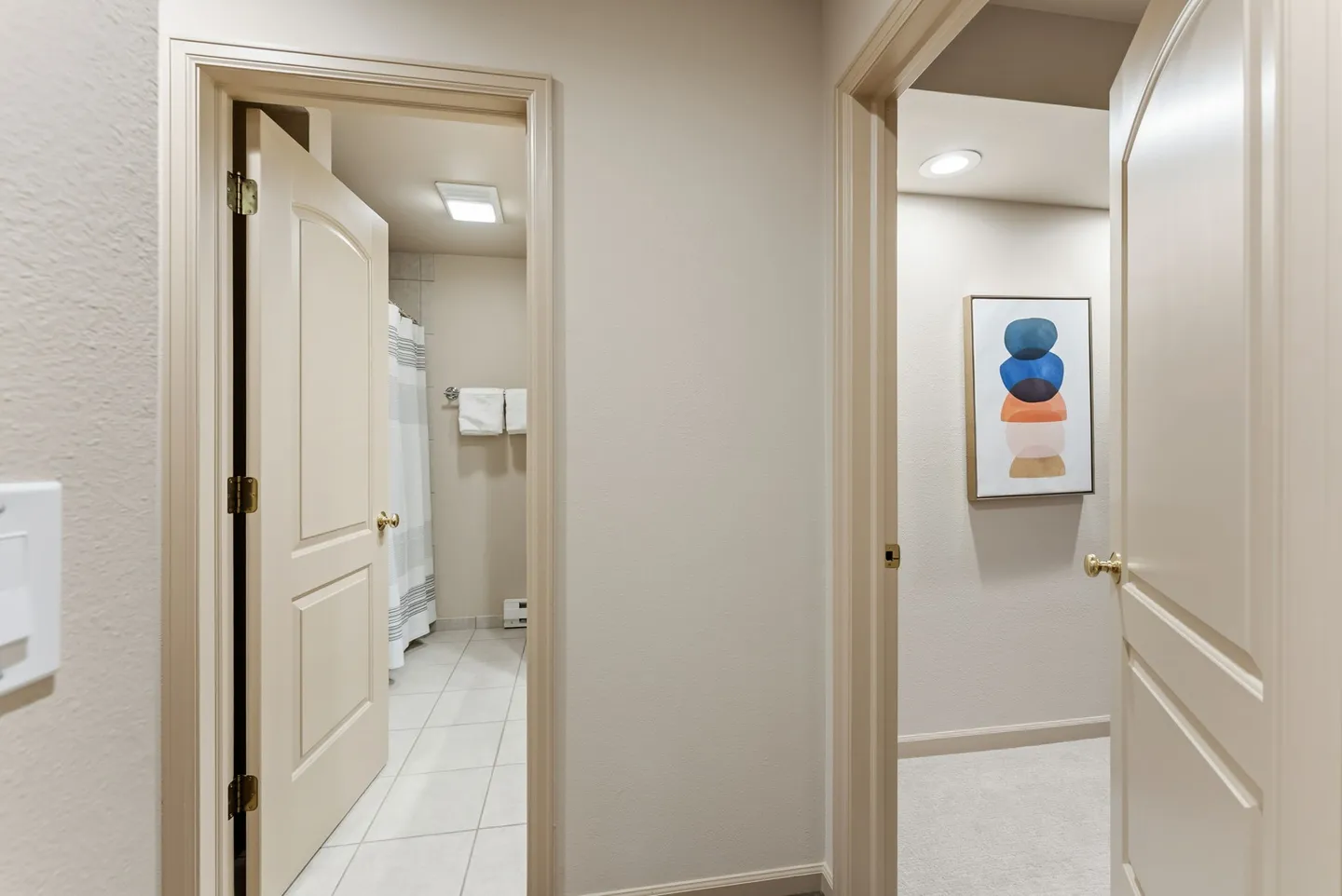 Hallway view into an open bathroom with tile flooring and an adjacent room with framed abstract art on the wall.