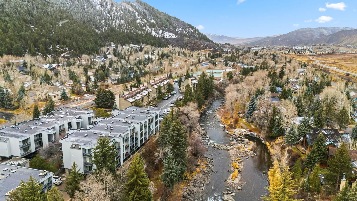 Aerial view of a river winding through a town with apartment buildings and snow-dusted mountains in the background.