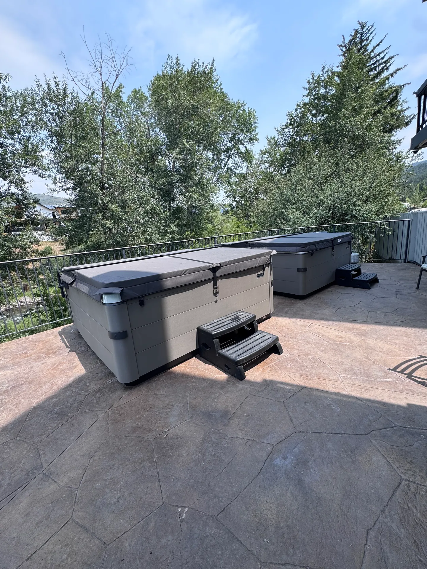 Outdoor patio with two covered hot tubs surrounded by metal railings and lush green trees.