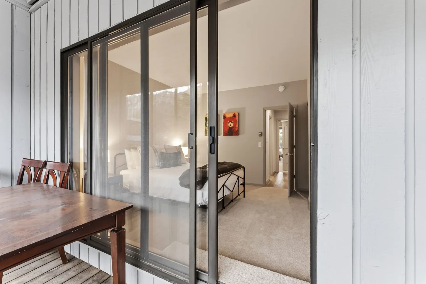 View through sliding glass doors from an outdoor patio into a carpeted bedroom with a made bed and bear artwork.
