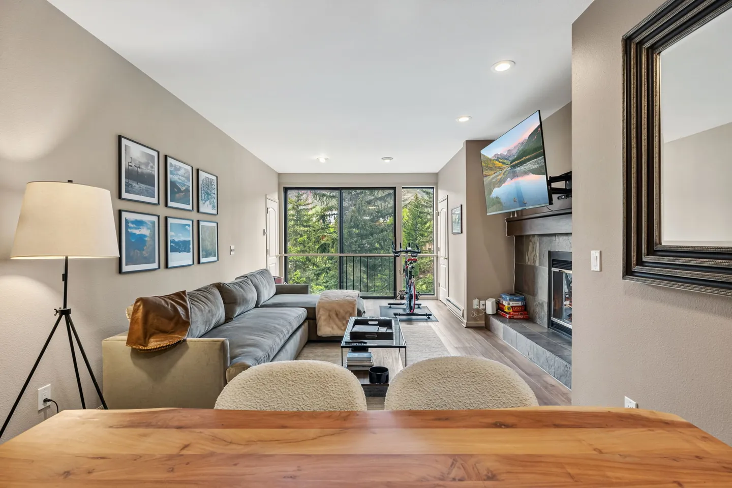 Living room featuring a sectional sofa, coffee table, wall-mounted TV over a fireplace, exercise bike, and sliding glass doors to a balcony with forest views.