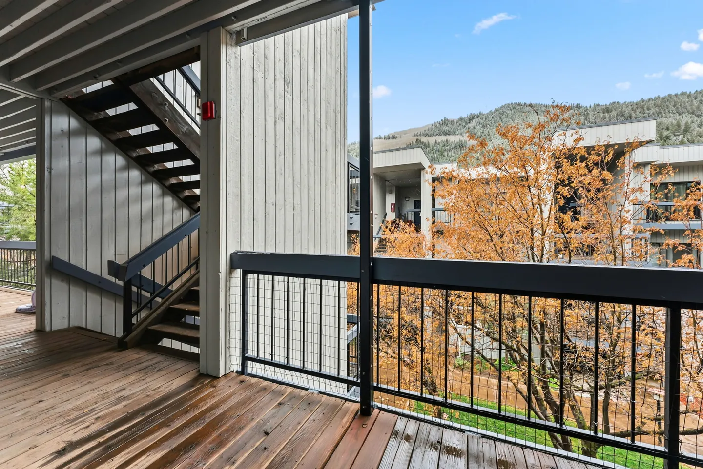 Covered outdoor walkway and stairs with wood plank flooring overlooking courtyard buildings and a hillside.