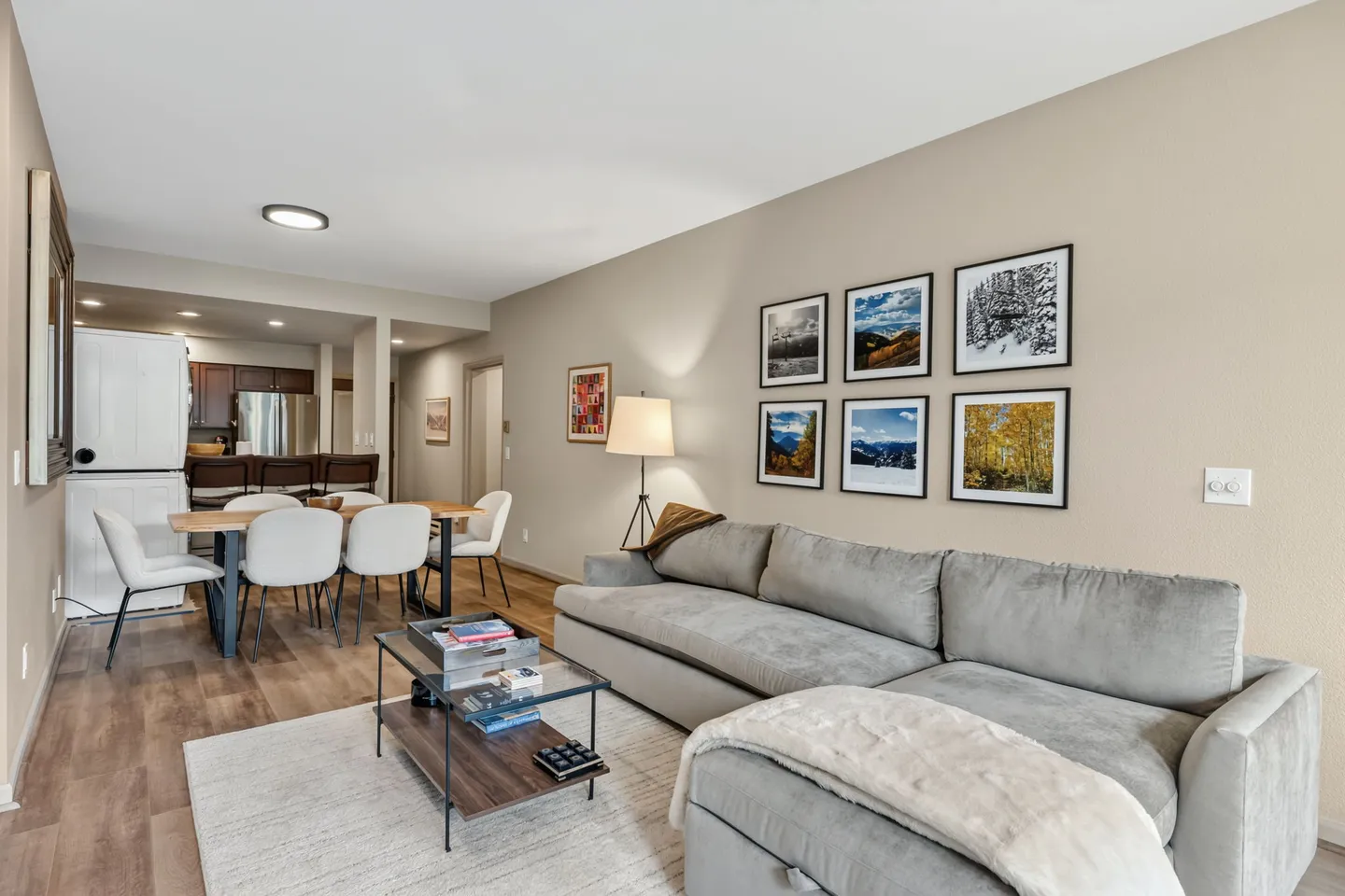 Open-concept living room with a gray sectional sofa, coffee table, gallery wall of nature photos, dining table, and view into the kitchen.