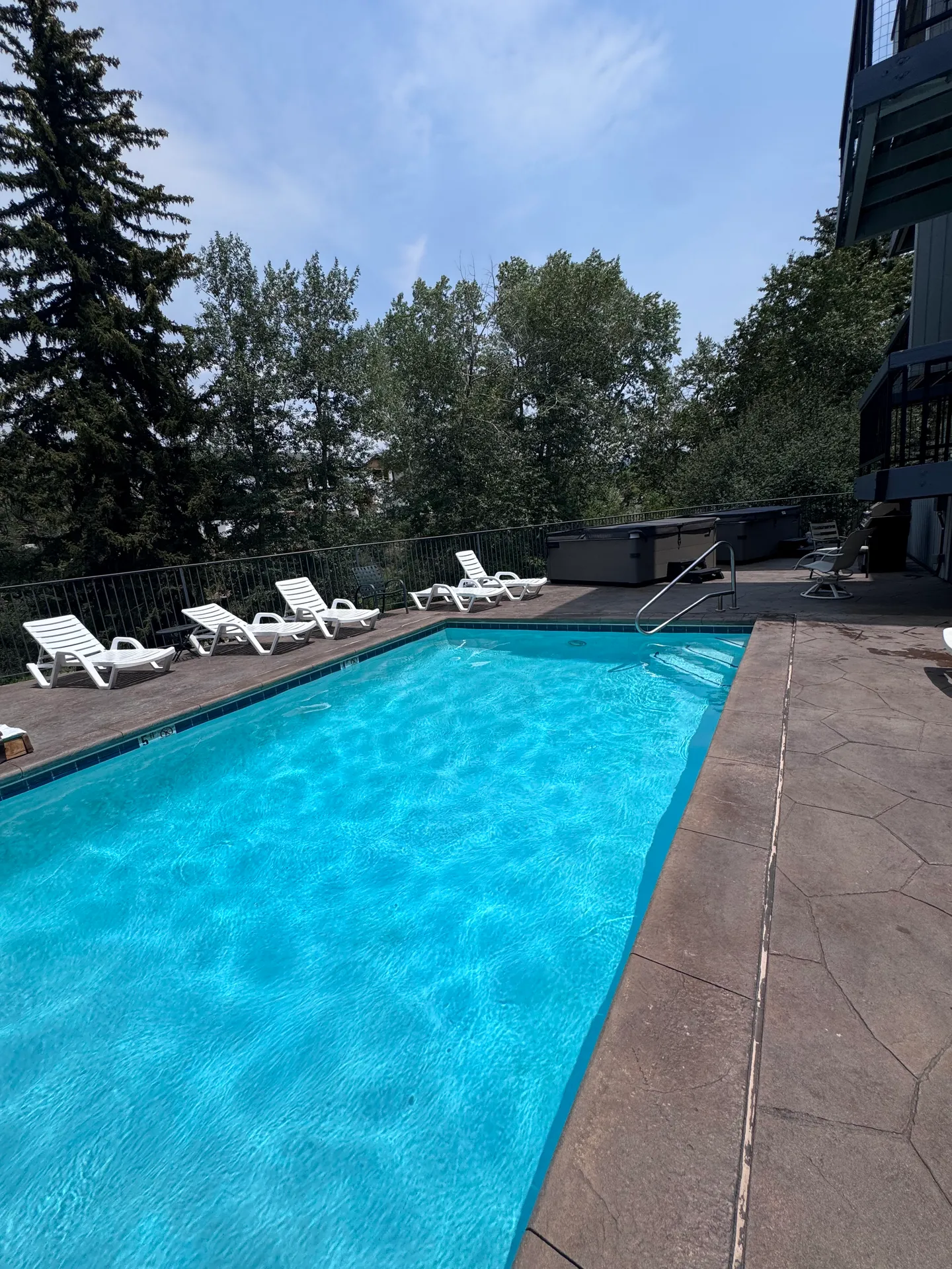 Outdoor swimming pool surrounded by a stone-look deck, white lounge chairs, hot tubs, and green trees under a blue sky.