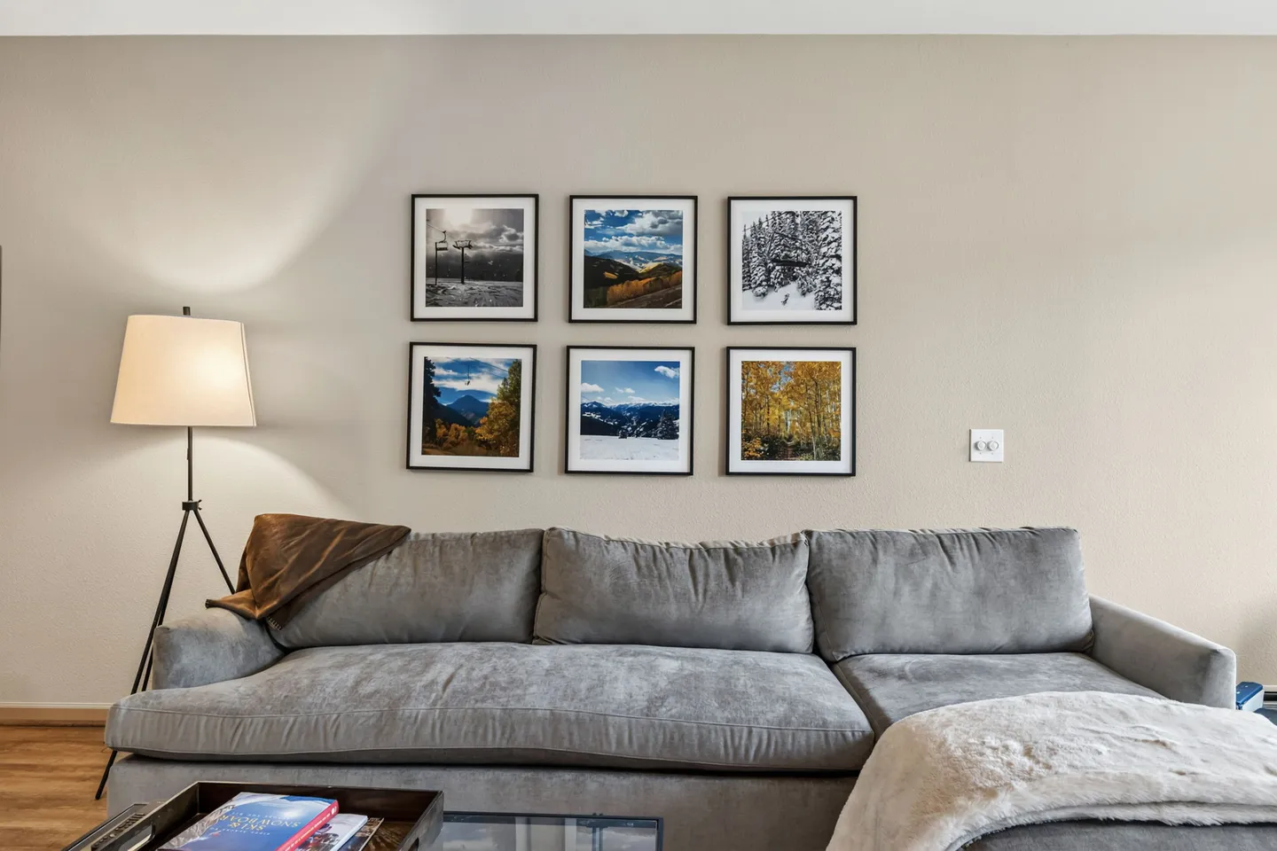Gray sectional sofa with a tripod floor lamp and a grid of six framed mountain landscape photographs on the wall.