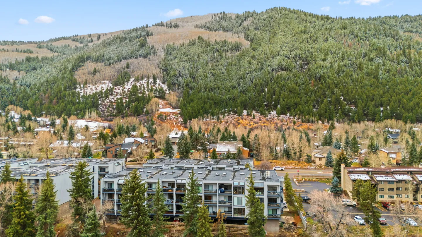 Aerial view of a multistory residential complex set against a large, tree-covered mountain slope with patches of snow.