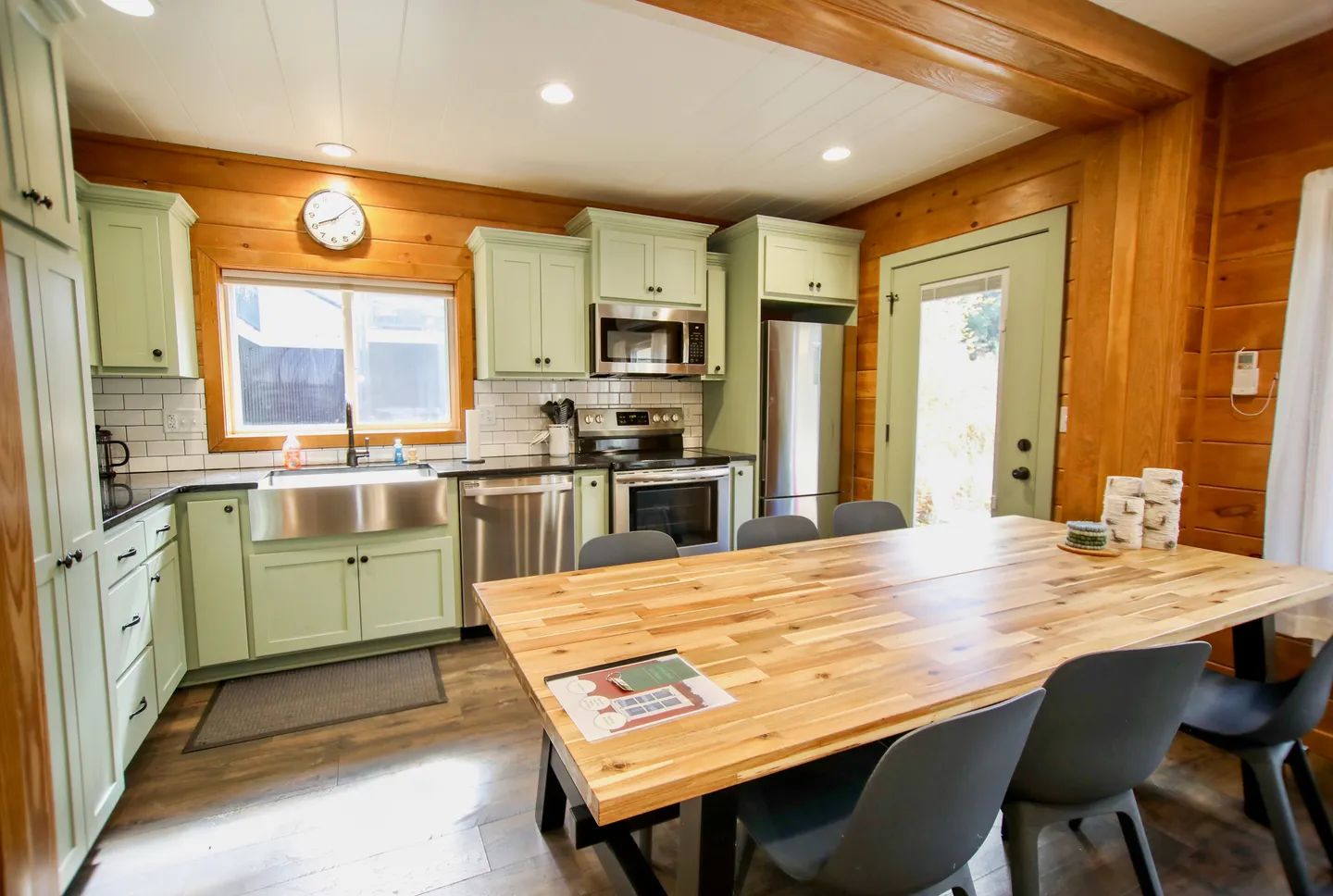 Kitchen with light green cabinets, stainless steel appliances, wood-paneled walls, and a large dining table with gray chairs.