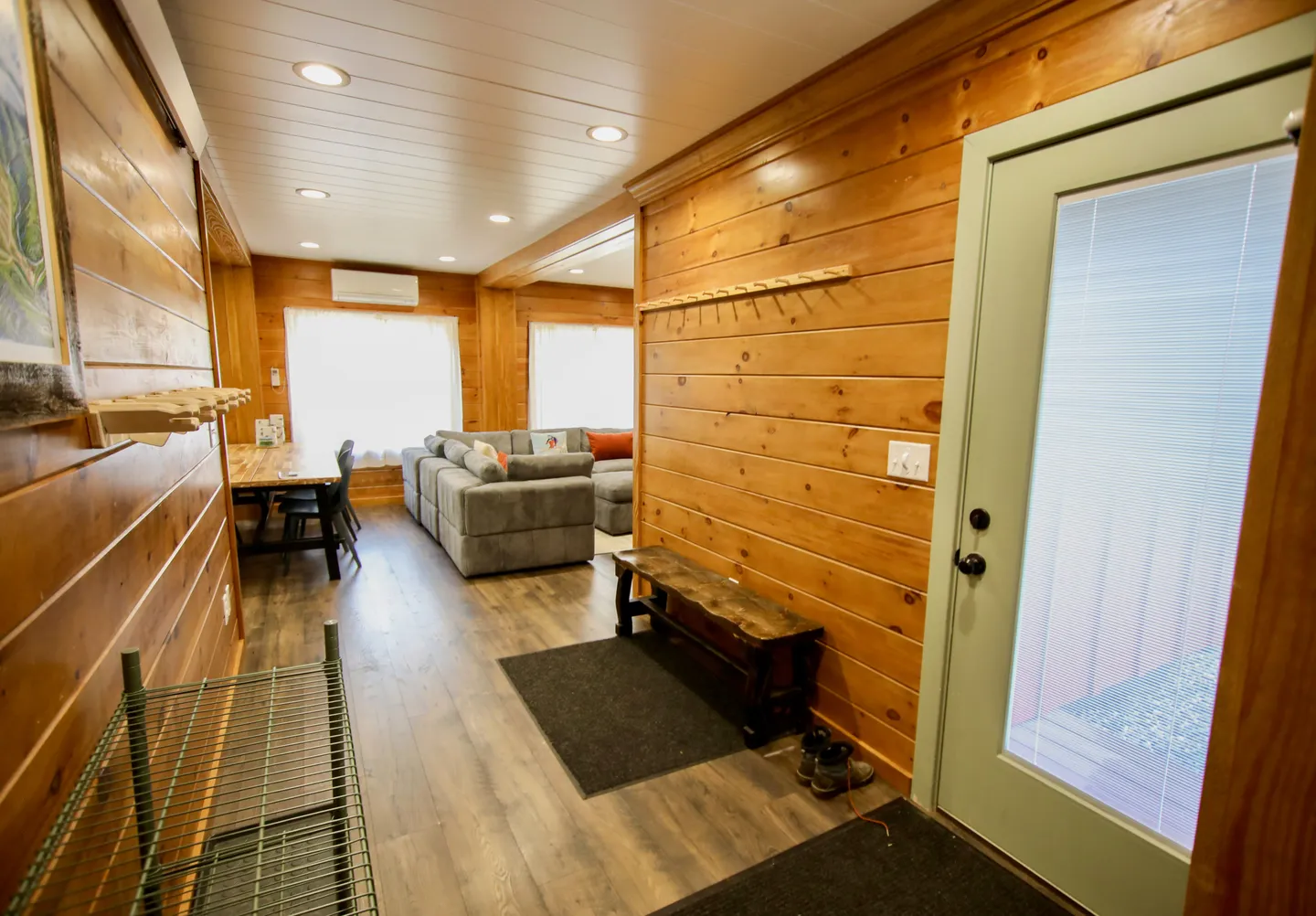 Entryway with wood-paneled walls, a small bench, and wire shelving, leading into an open living room with a sectional sofa.