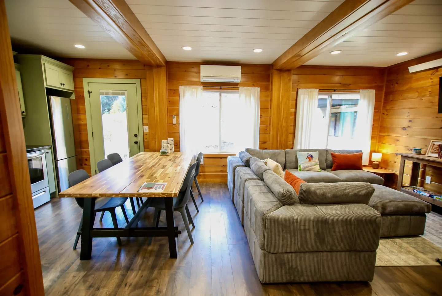 Open concept living room and dining area with wood-paneled walls, exposed ceiling beams, a large sectional couch, and a dining table.