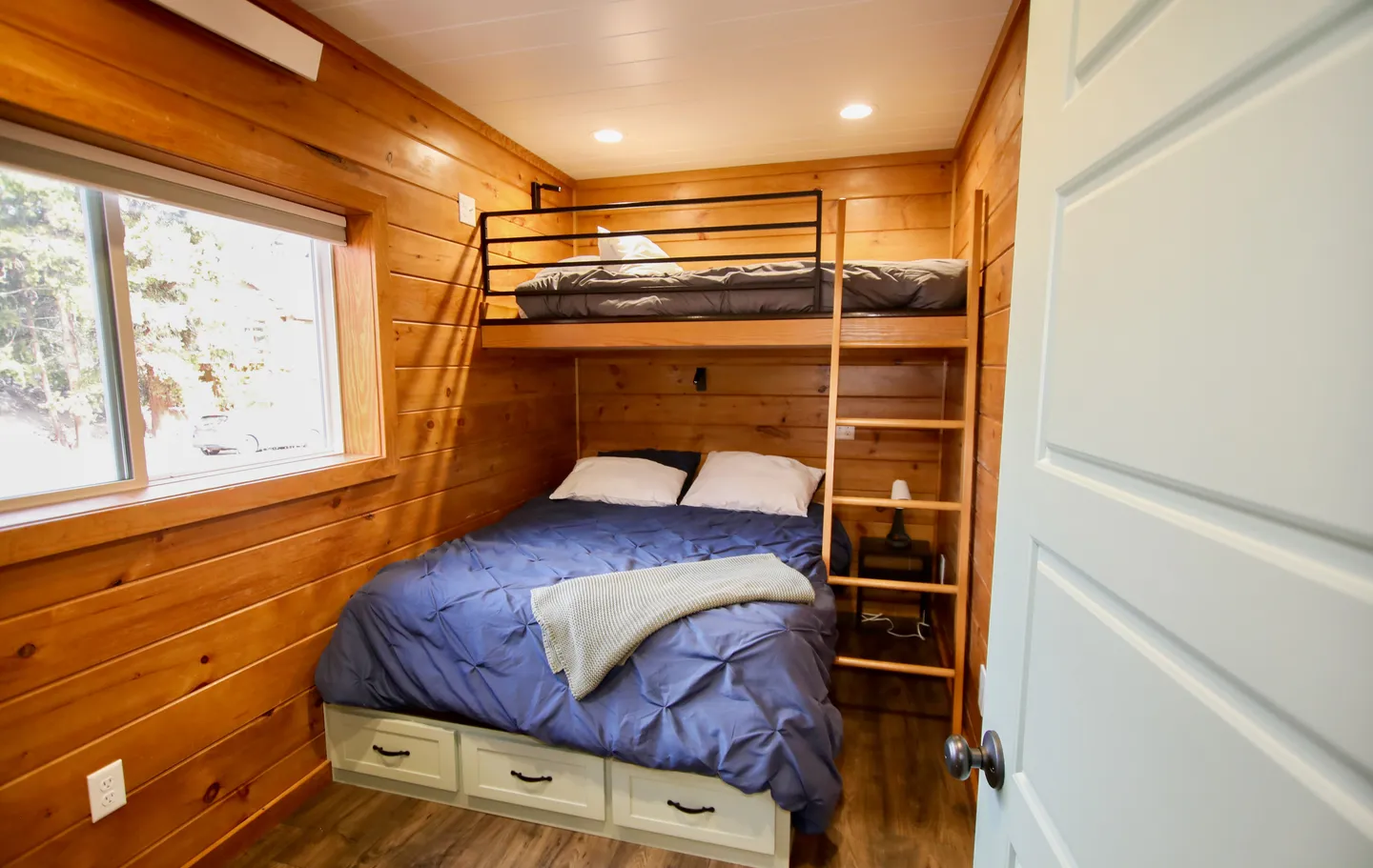 Bedroom with wood-paneled walls featuring a lower bed with drawers beneath and an upper bunk with a wooden ladder.