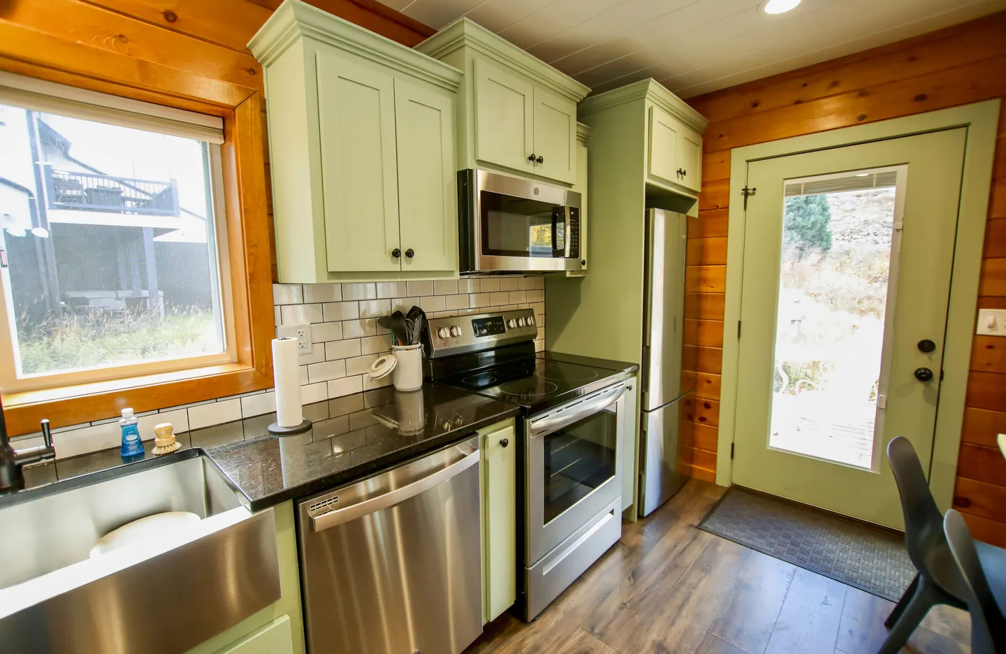 Kitchen with sage green cabinets, stainless steel appliances, dark countertops, white subway tile backsplash, and a log-style wood wall.