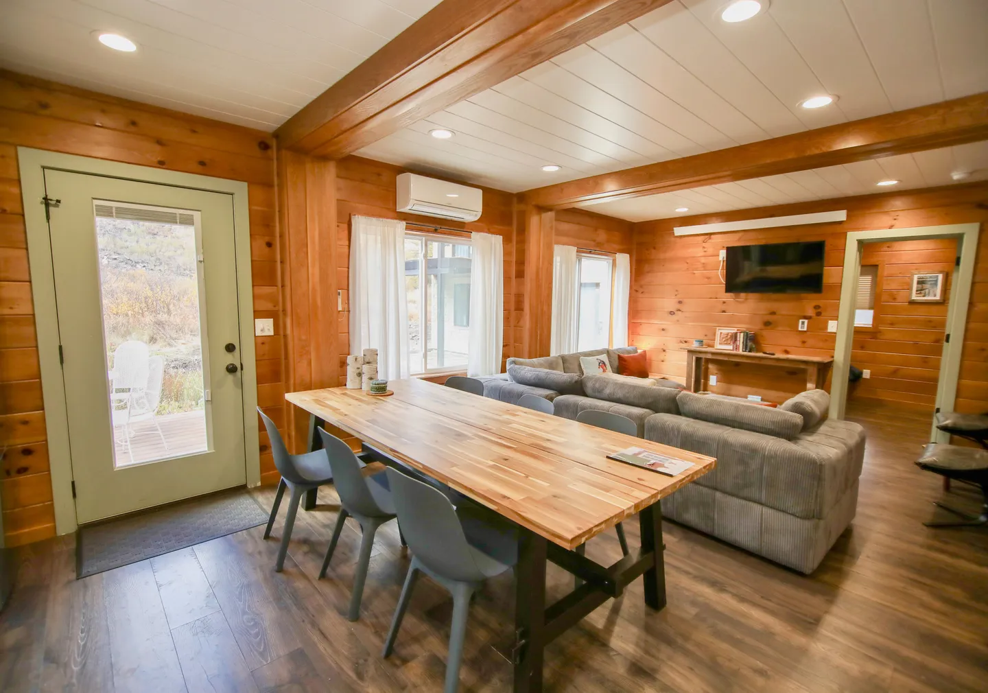 Open-concept cabin interior featuring a wood dining table with chairs, an adjacent living area with a sectional sofa and wall-mounted TV, and log-style wood walls.