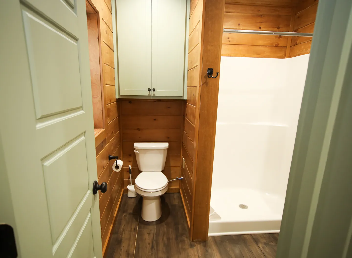 Bathroom with wood-paneled walls, a toilet, an overhead mint green cabinet, and a walk-in shower stall with white surrounds.