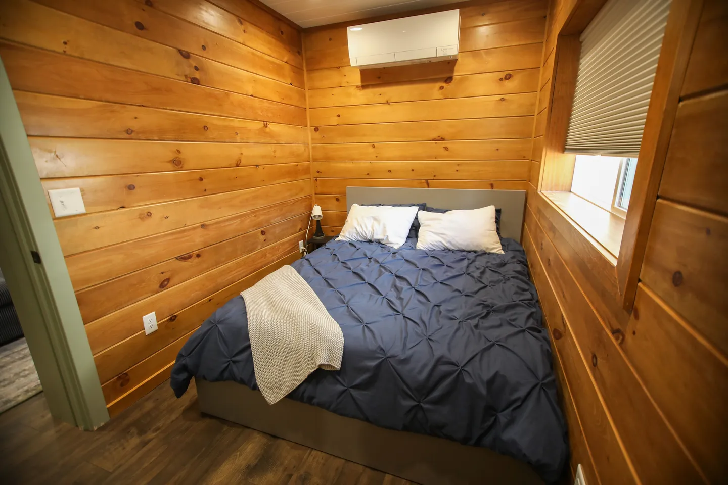 Bedroom with wooden plank walls, a bed dressed in a dark blue comforter, and a wall-mounted climate unit.
