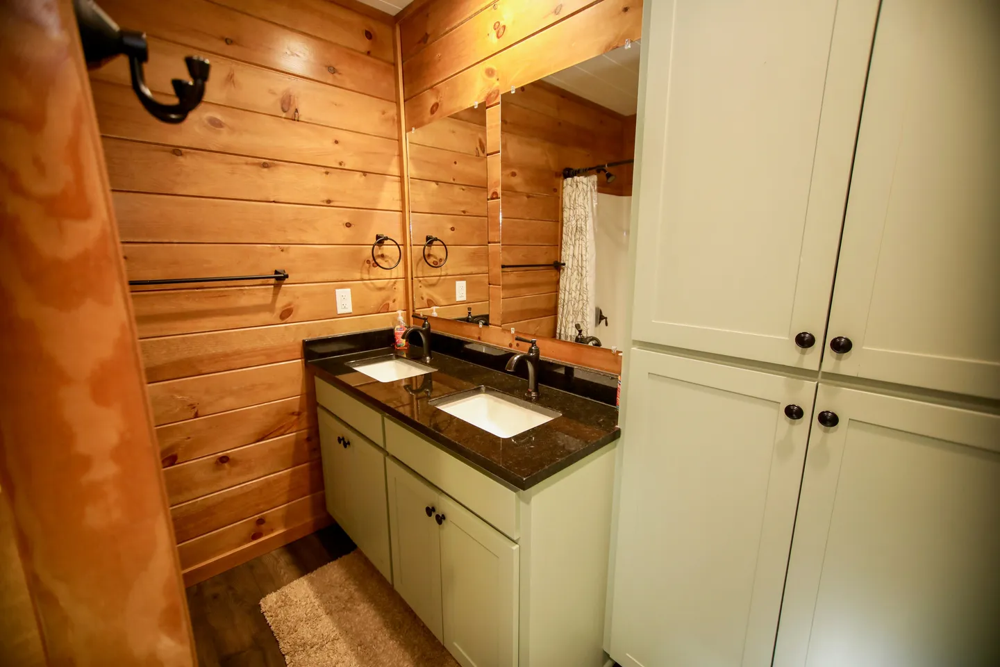 Bathroom with wood-paneled walls, a double vanity with dark countertops, and tall light-green storage cabinets.