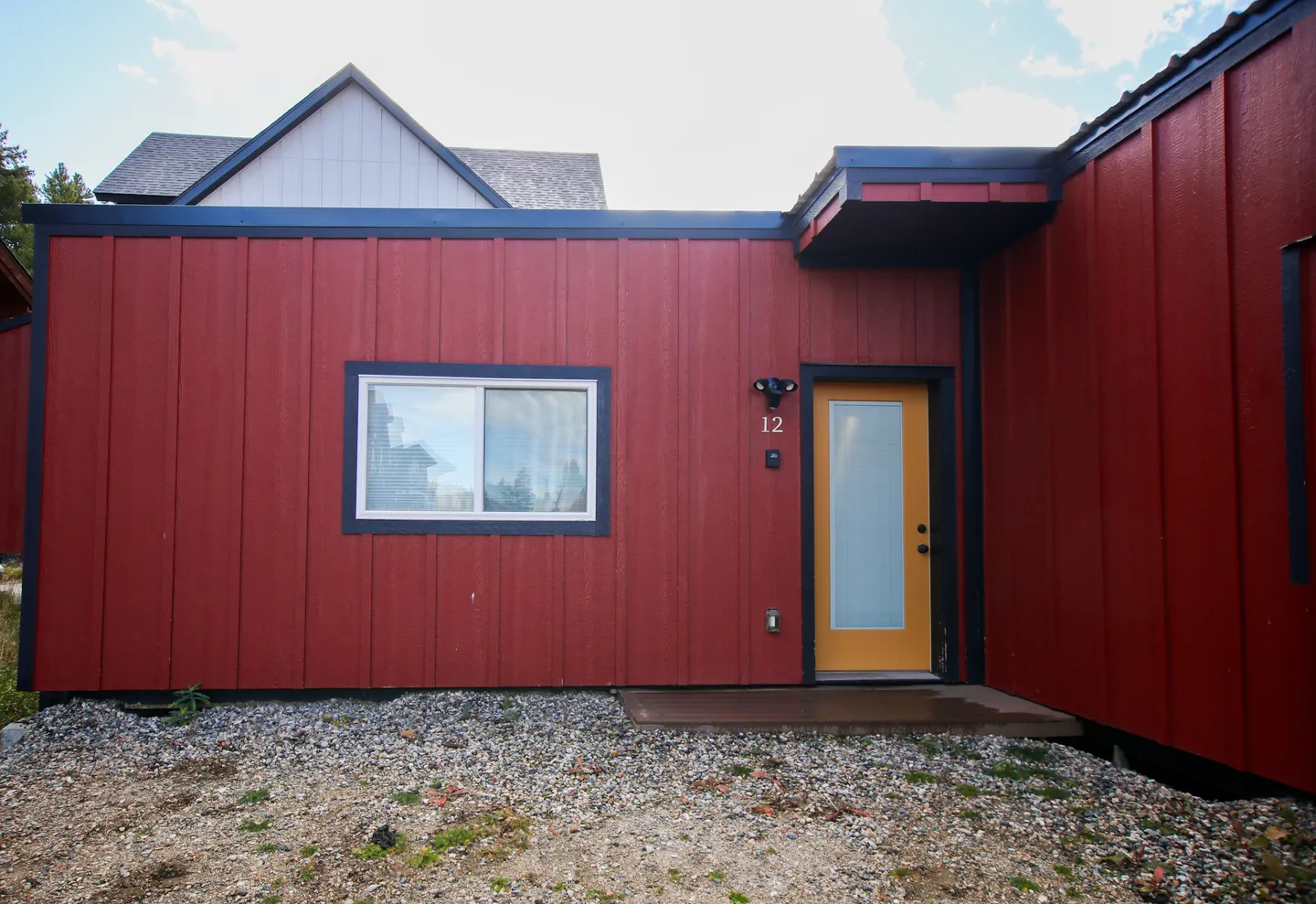 Exterior entrance of a house with red vertical siding, a yellow front door, and gravel ground in front.