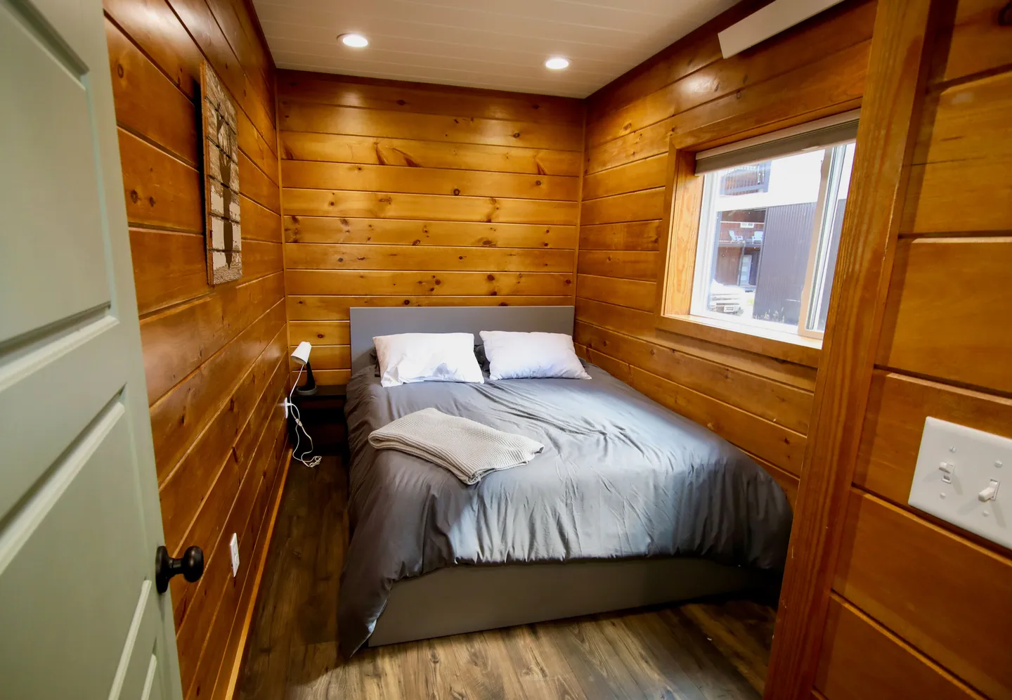 Bedroom with wood-paneled walls, wood-look flooring, and a bed with grey bedding fitting into an alcove beneath a window.