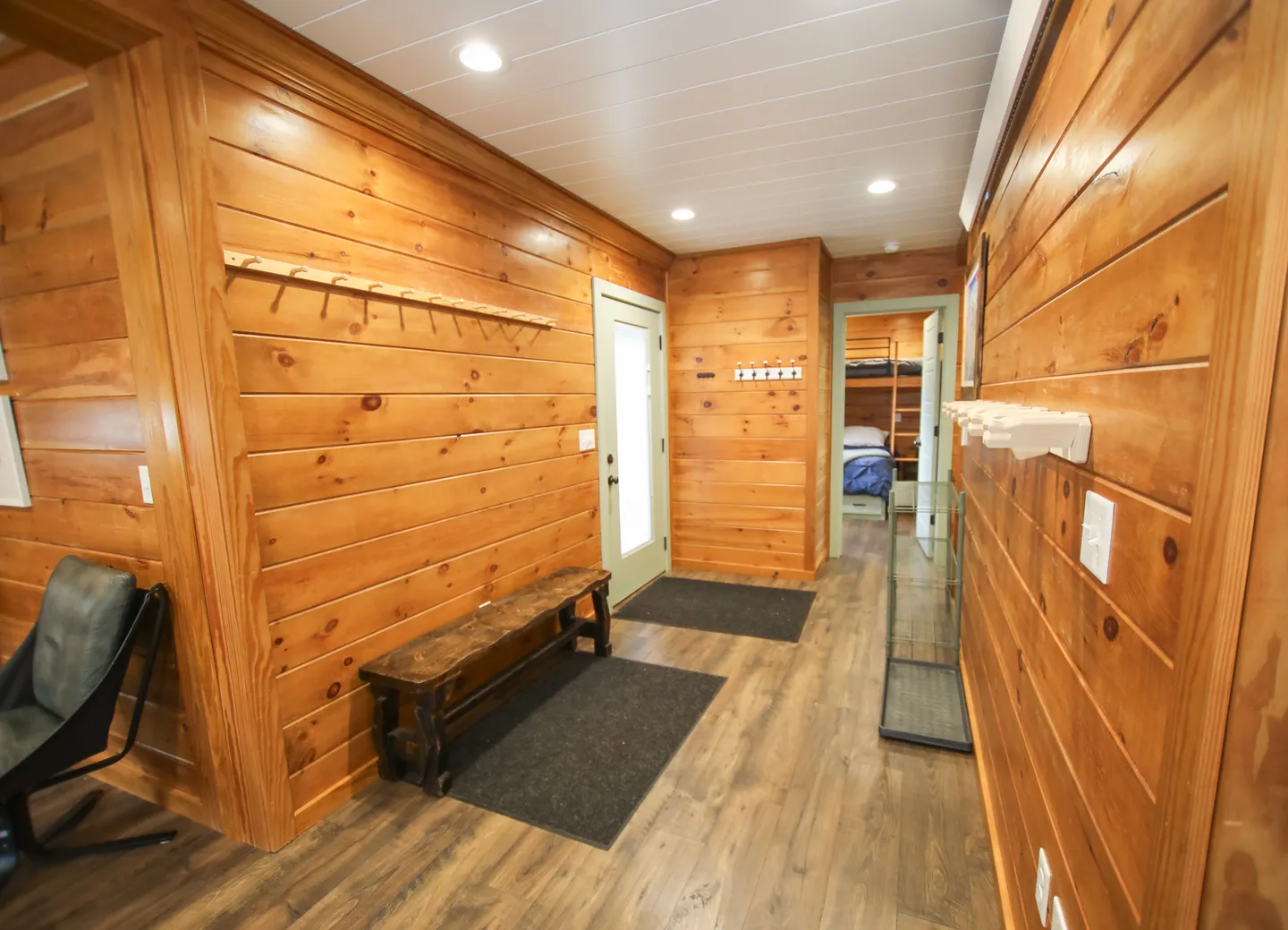 Entryway hallway with pine-paneled wood walls, wood-look flooring, a wooden bench, wall-mounted coat racks, and a doorway leading into a bunk bedroom.