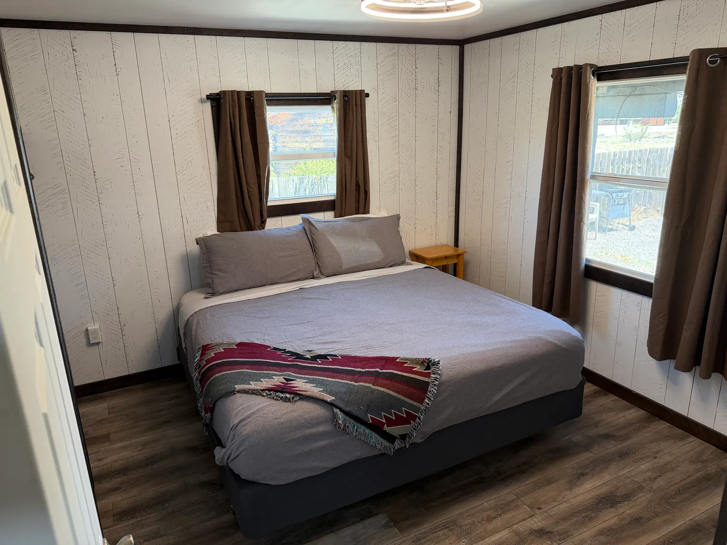 Bedroom with a made bed, wood-plank style walls, wood-look flooring, and two windows with brown curtains.