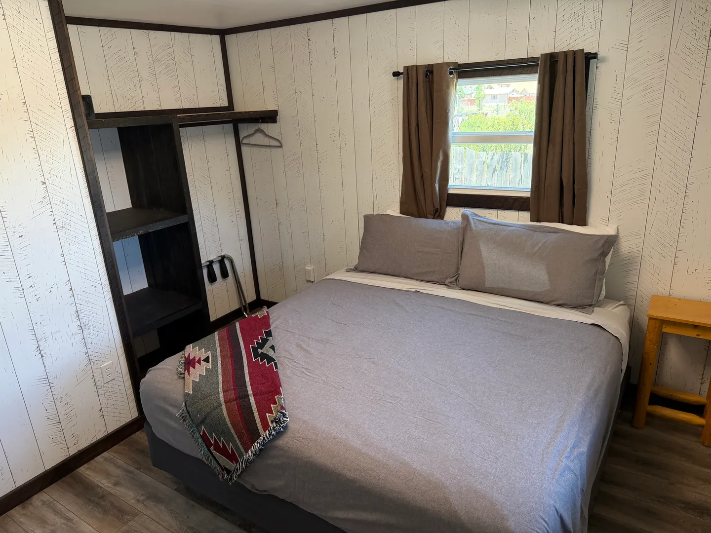 Bedroom with a bed made in grey linens, wood-paneled walls, an open dark wood closet organizer, and a window with brown curtains.