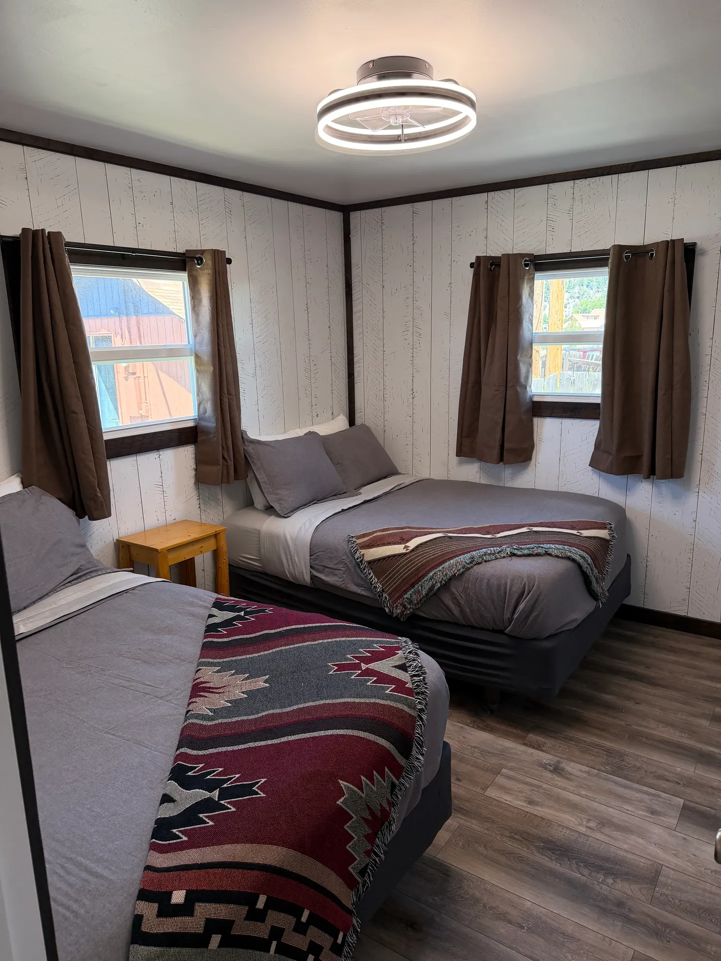 Bedroom with two beds, patterned blankets, wood-look flooring, and white paneled walls with two curtained windows.