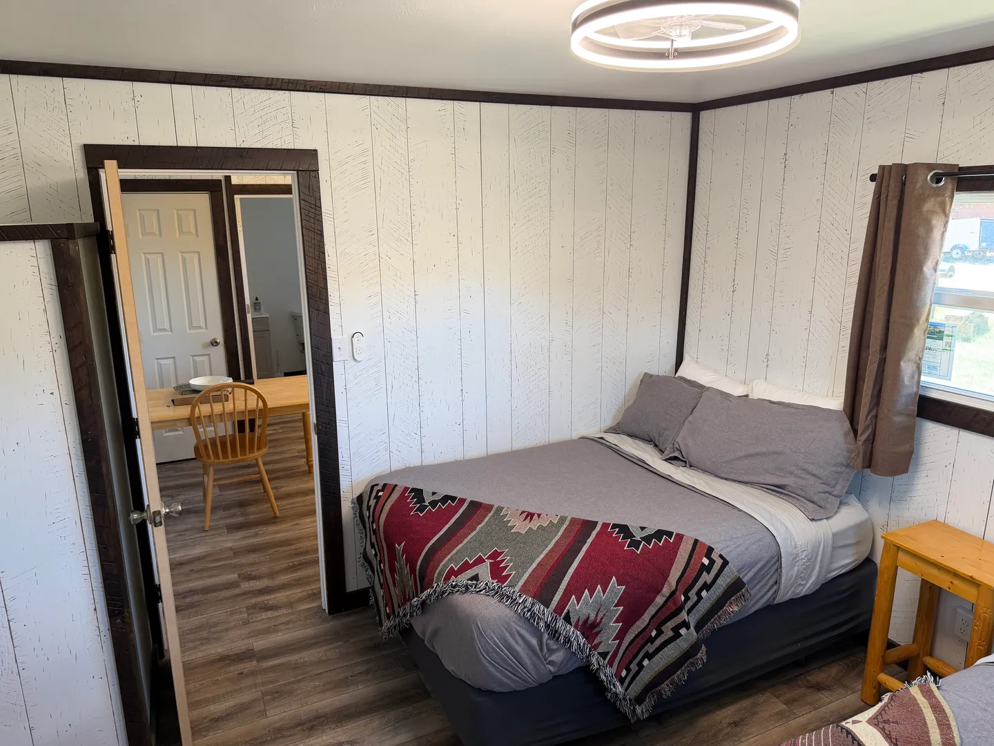 Bedroom with white vertical wood-paneled walls, a made bed with grey linens and a patterned throw blanket, and an open doorway leading to a dining area.