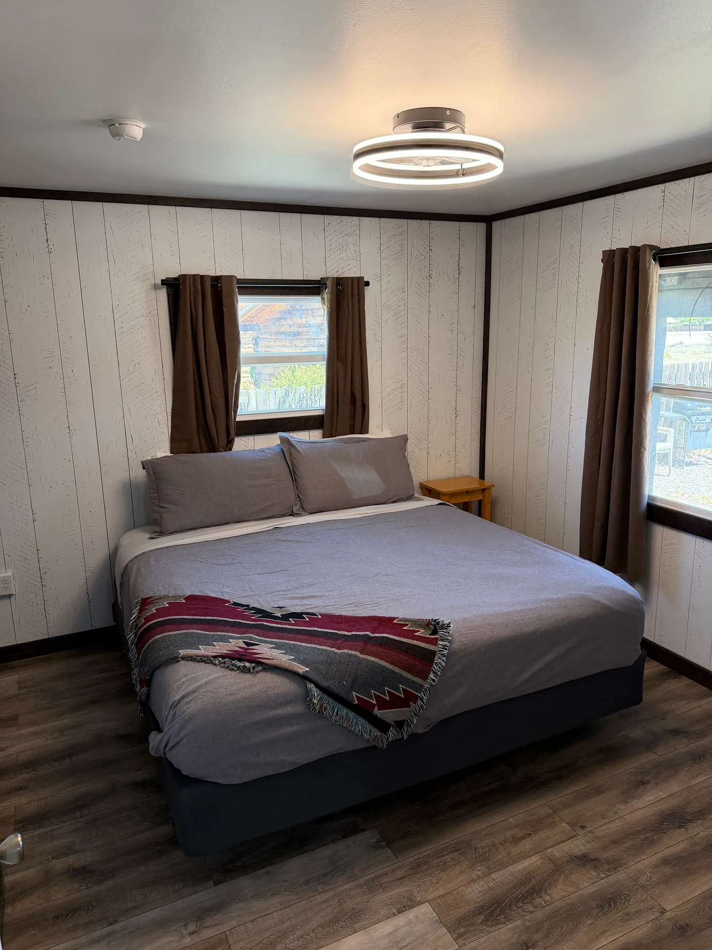 Bedroom with a made bed, wood-plank style accent walls, dark curtains on two windows, and a modern circular ceiling light.
