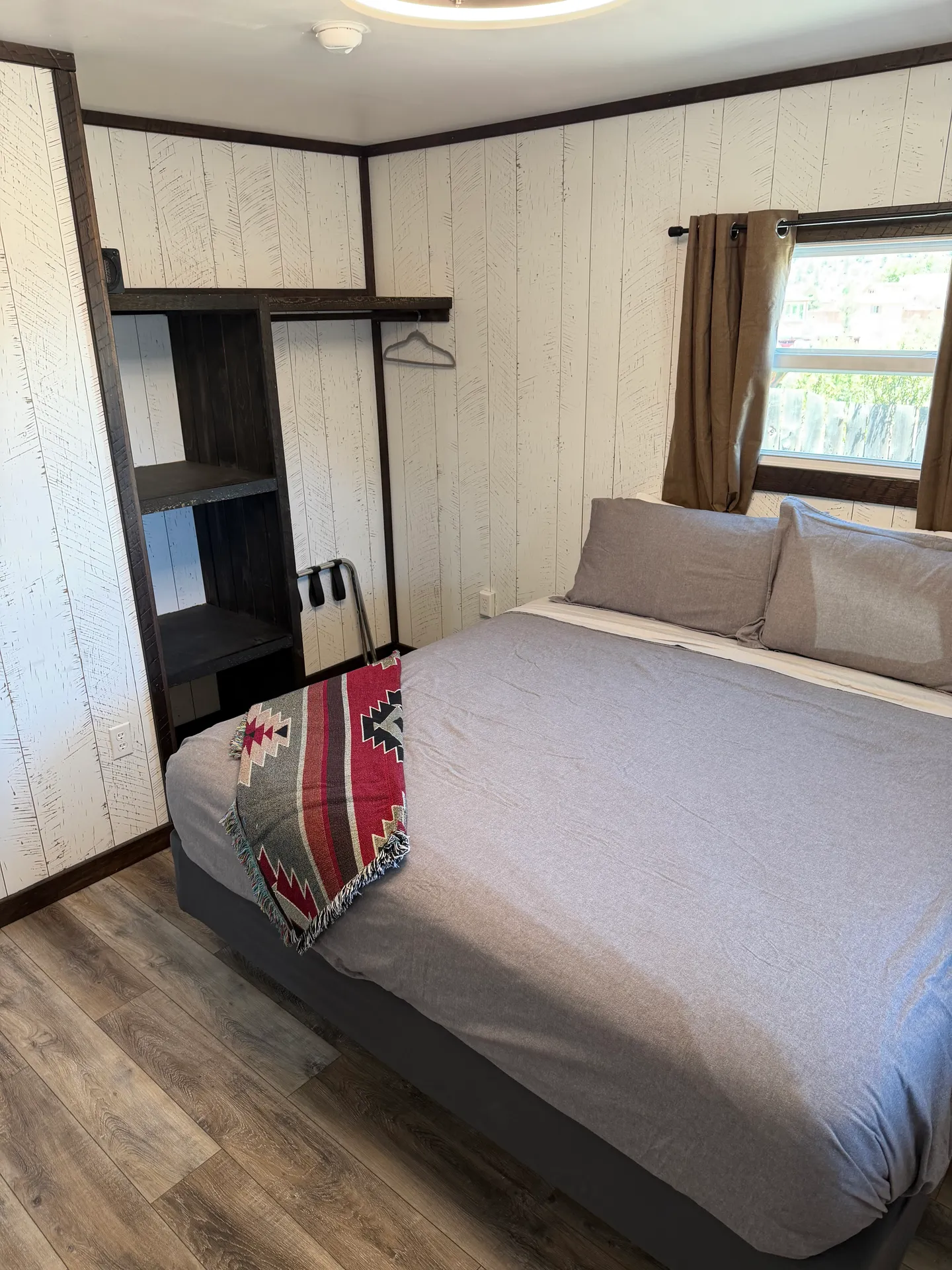 Bedroom with a bed made in gray linens, light wood-look panel walls, wood-style flooring, and an open dark wood shelving and hanging closet unit.