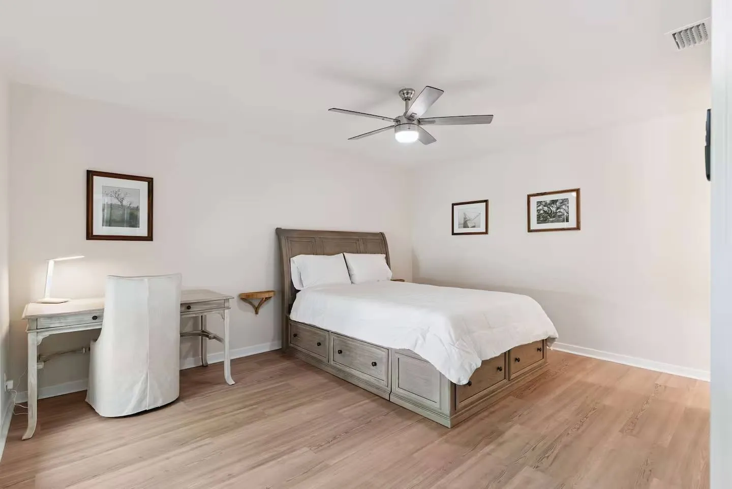 Bedroom featuring a wooden sleigh bed with built-in storage drawers, a writing desk with a white slipcovered chair, wood-style flooring, and a ceiling fan.