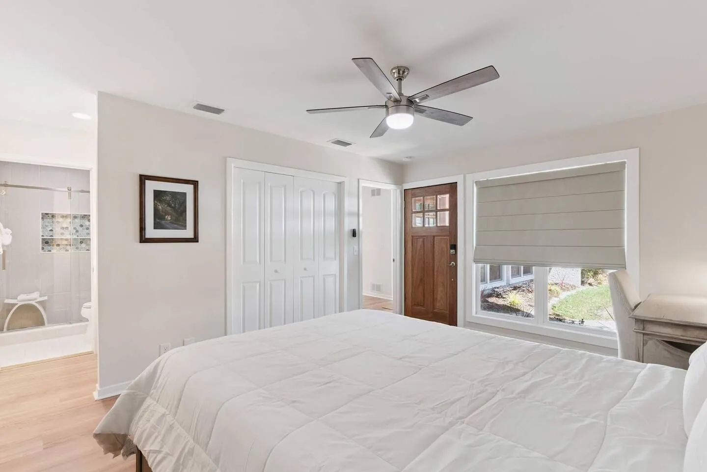 Bedroom with a white bed, ceiling fan, wooden exterior door, large windows, and an en-suite bathroom visible to the left.