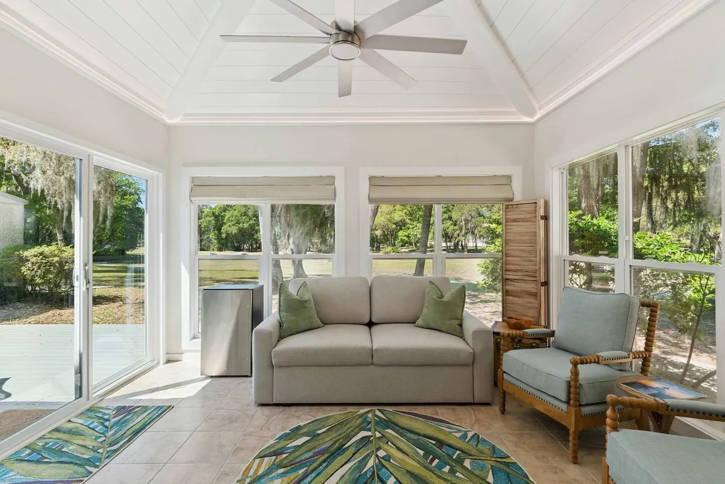 Sunroom with a vaulted white-paneled ceiling, large windows showing wooded views, a sofa, an armchair, and tile flooring.