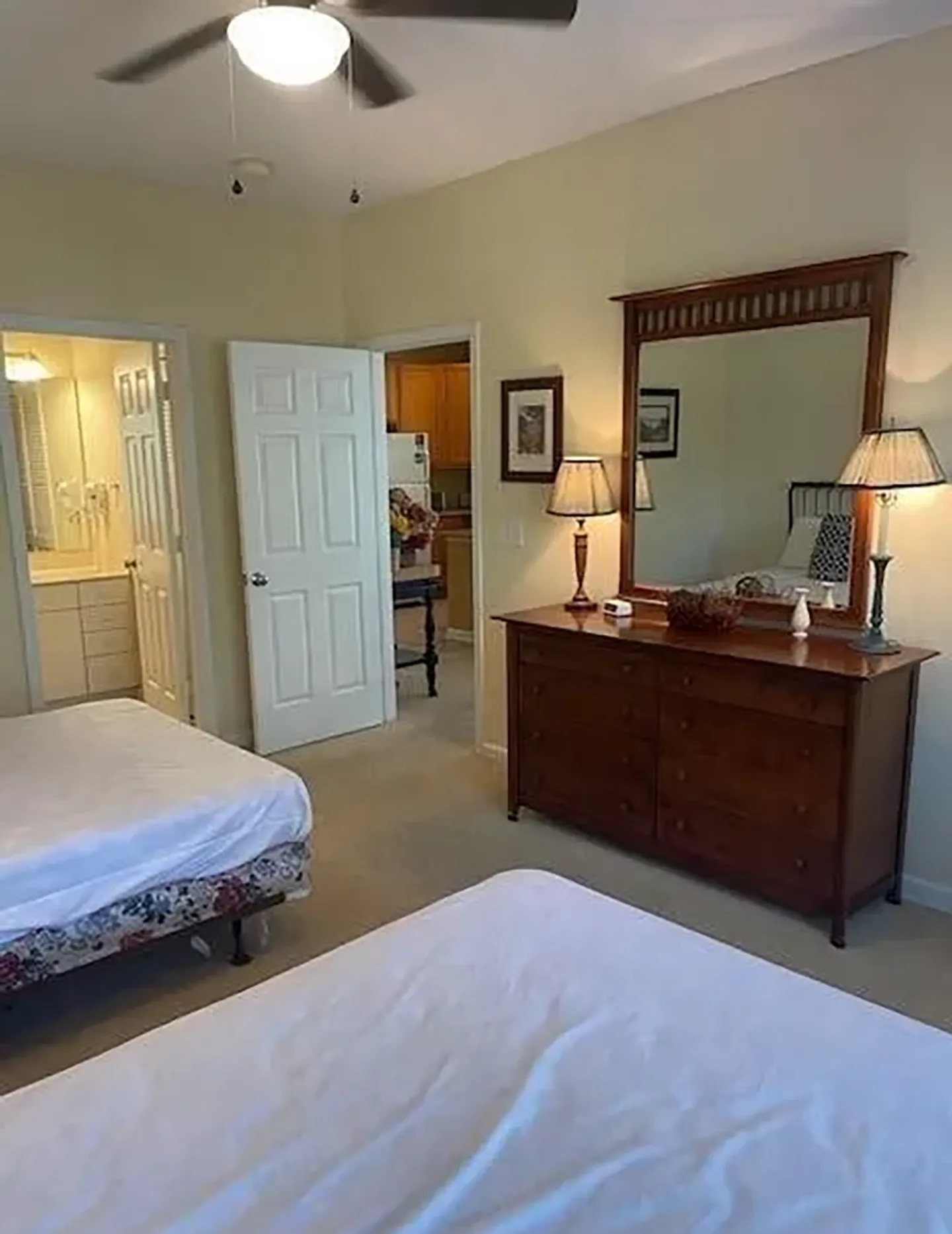 Bedroom with two beds, a wooden dresser with a large mirror, and open doors leading to a bathroom and kitchen area.