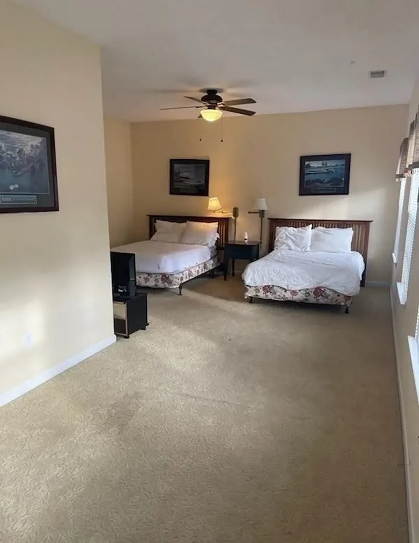 Bedroom with two beds, beige walls, wall-to-wall carpet, a ceiling fan, and framed artwork on the walls.