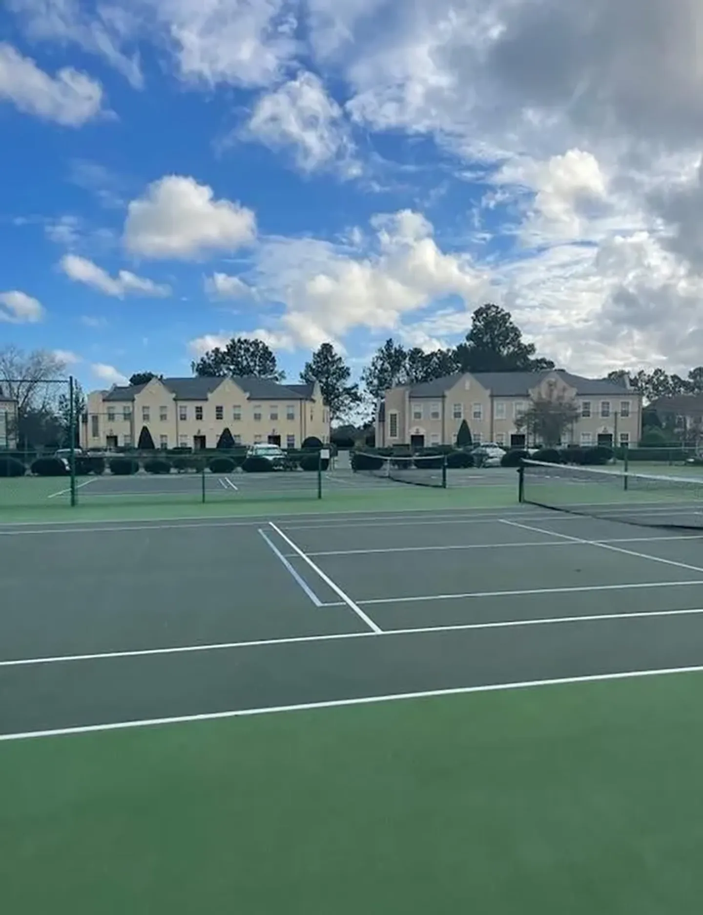 Outdoor tennis court in front of multi-story residential buildings under a partly cloudy sky.