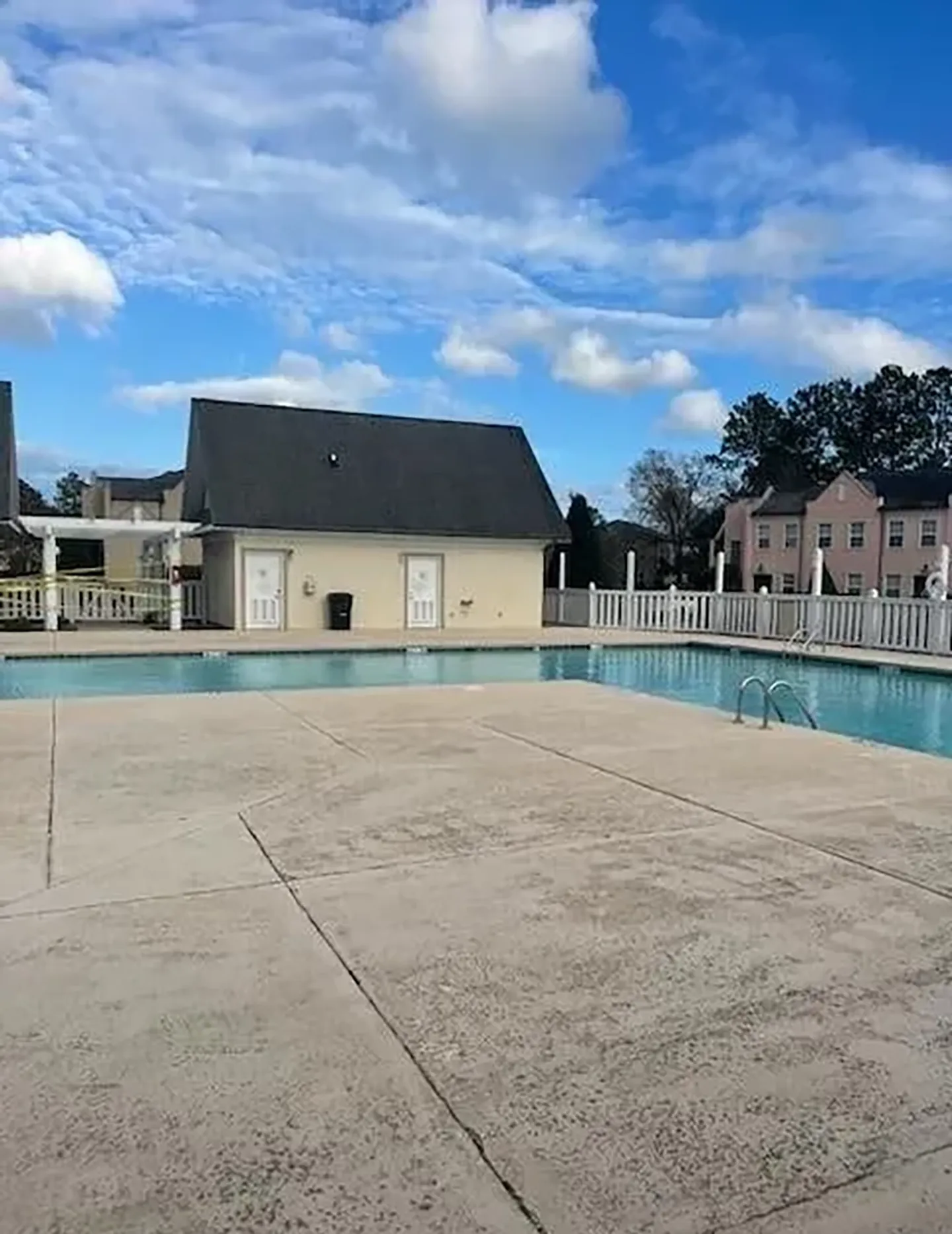 Outdoor swimming pool surrounded by concrete decking and white fencing with a small pool house and townhomes nearby under a blue sky.