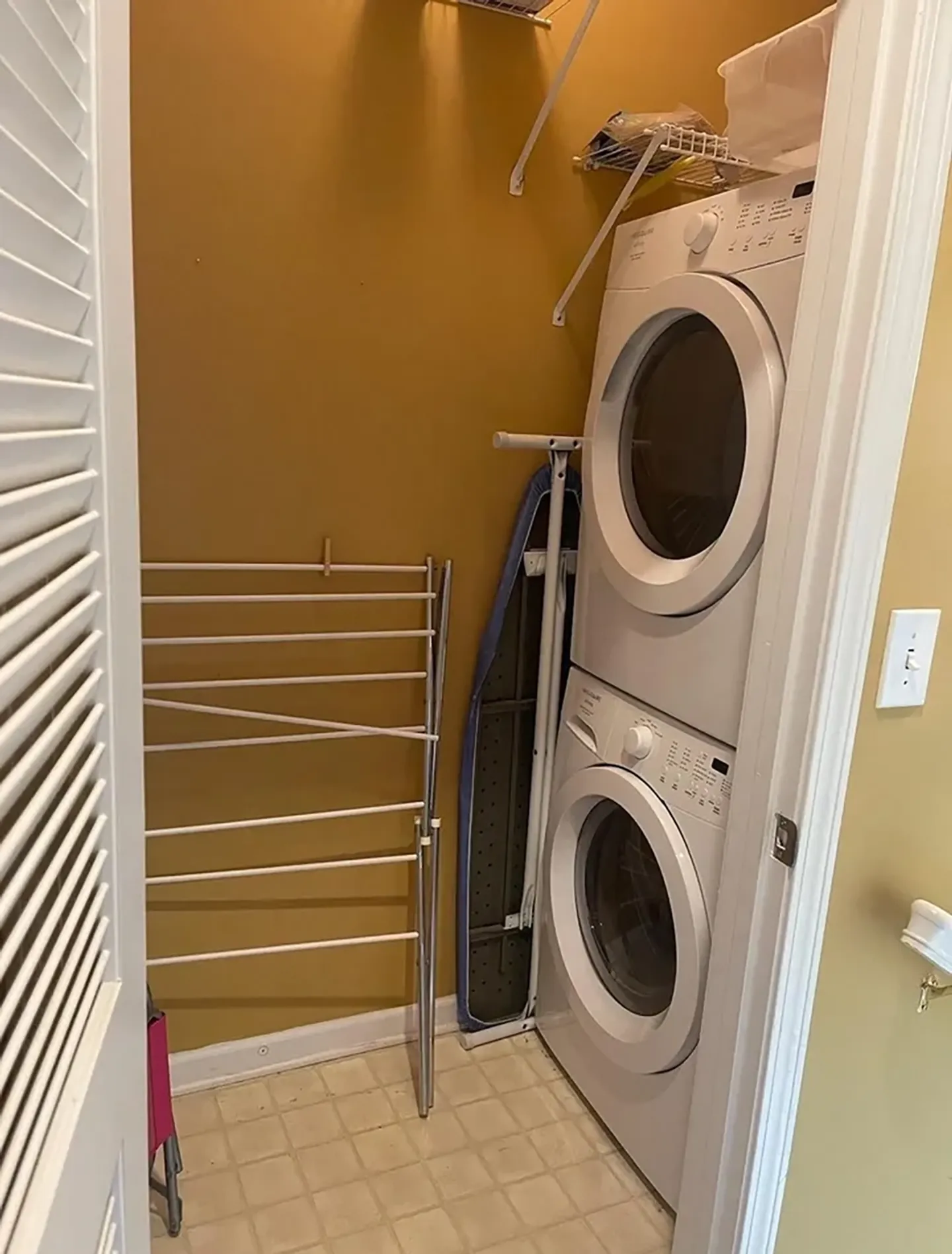 A laundry closet featuring a stacked white washer and dryer, a drying rack, and an ironing board.