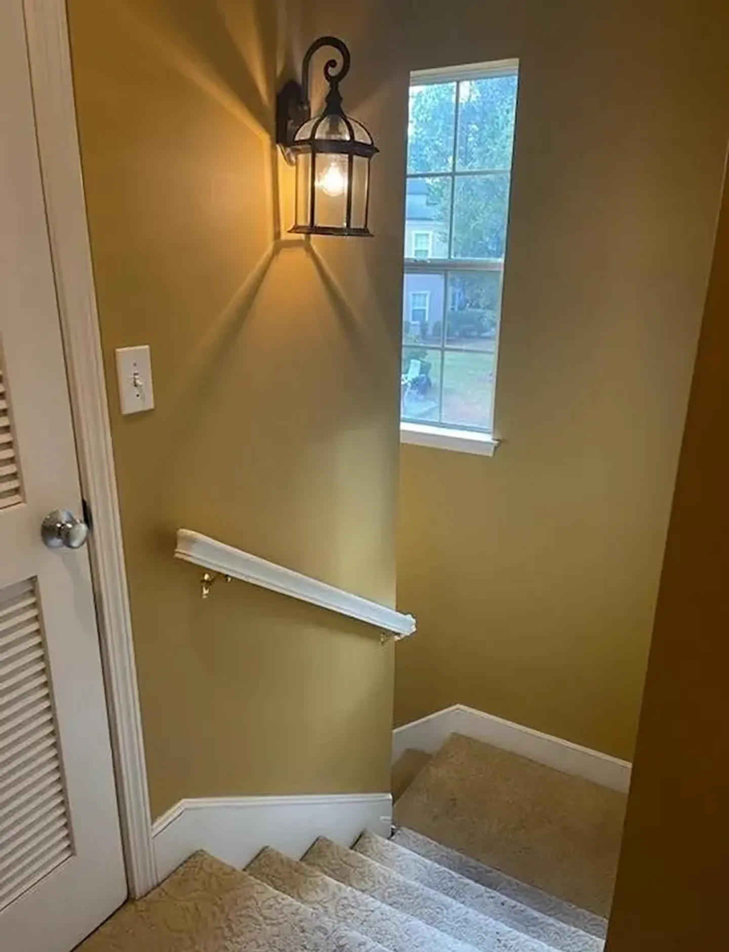 A carpeted staircase leading downwards with mustard yellow walls, a white railing, a lantern-style wall sconce, and a window.