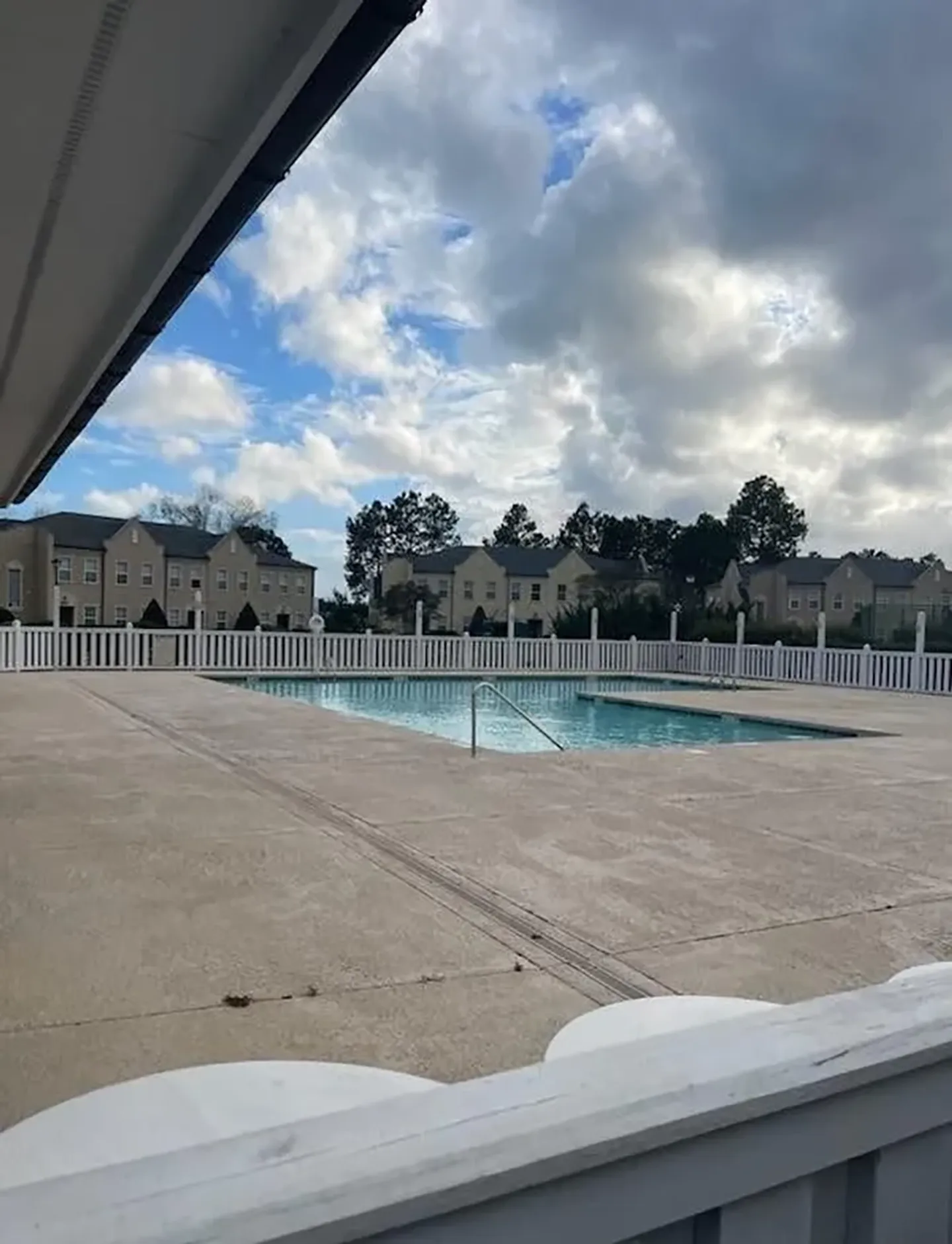 Outdoor fenced swimming pool with concrete deck surrounded by residential buildings under a cloudy sky.