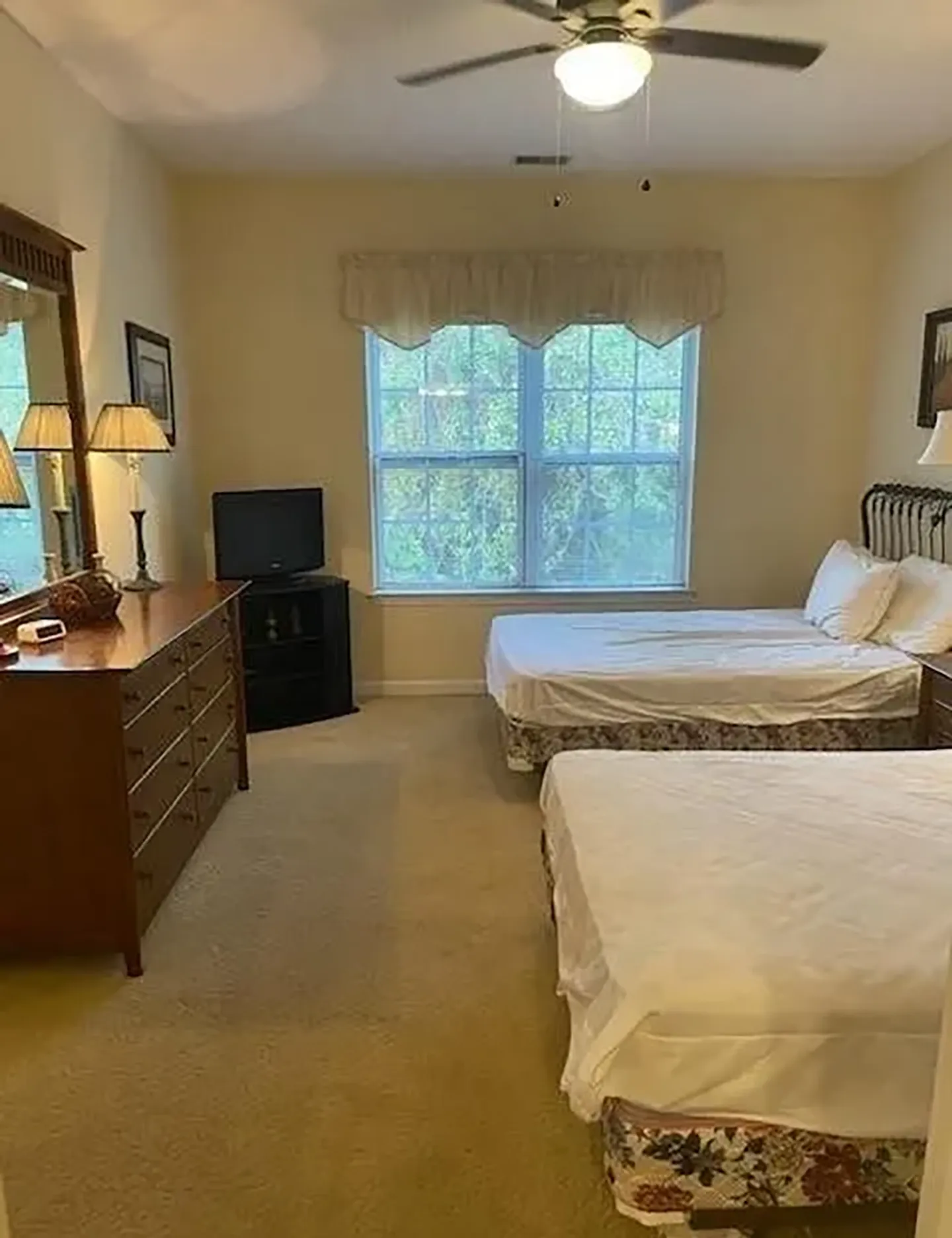 Bedroom with two beds, carpeted floor, wooden dresser, television in the corner, and a window with a valance.