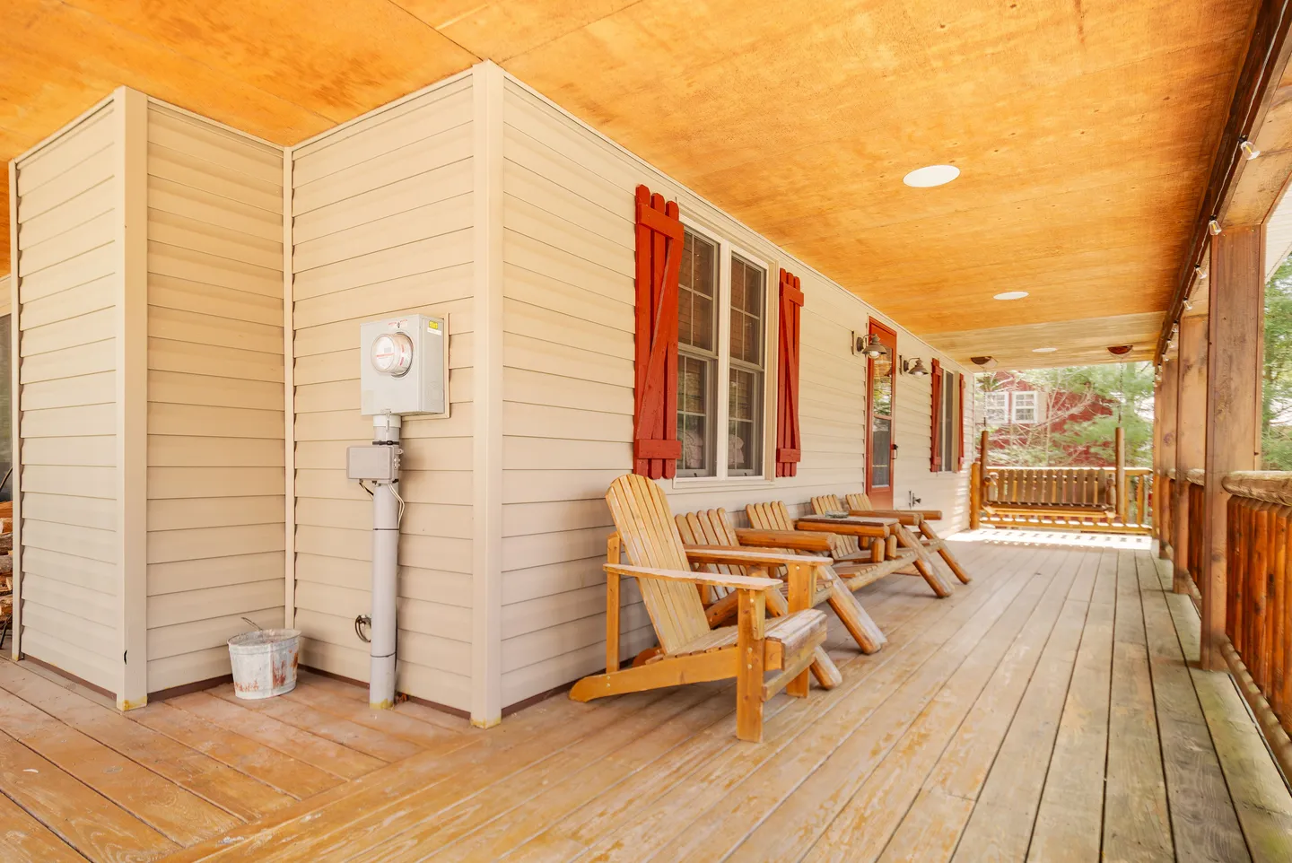 A long covered porch with wooden plank flooring, wooden armchairs, picnic-style seating, and red window shutters.