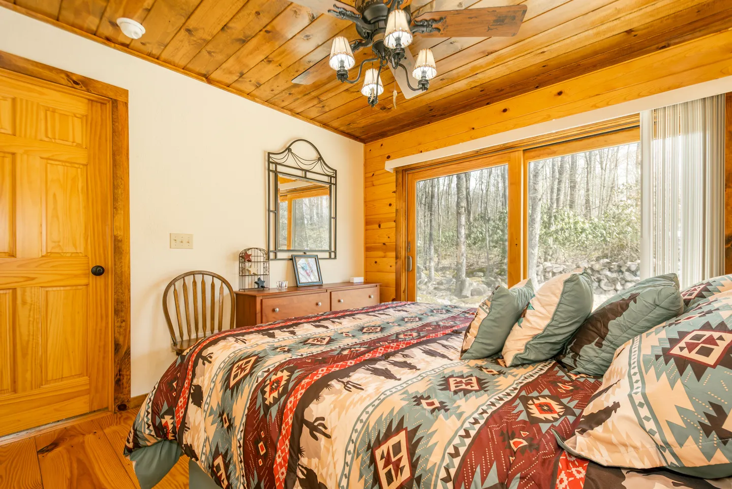 Bedroom with wood-paneled ceiling, rustic patterned bedding, and sliding glass doors looking out to trees.